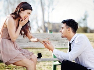  A man wearing a white shirt, blank pants and black tie proposes to a woman down on one knee to a woman wearing a pink sundress. 