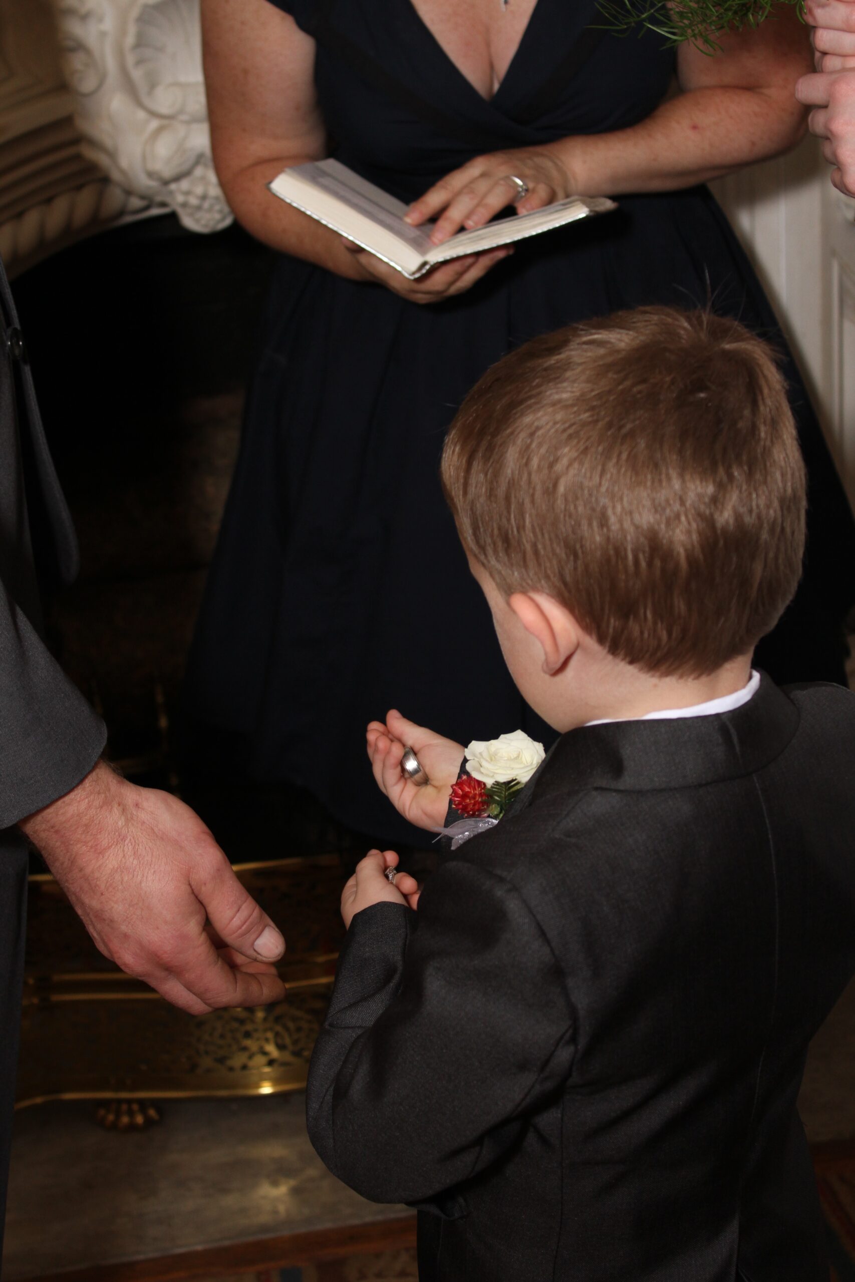 The groom's hand reaches down for the rings as the little ring bearers reaches out with one ring in each tiny fist. The ring bearer is facing away from the camera wearing a navy suit with a red and white boutonniere. 