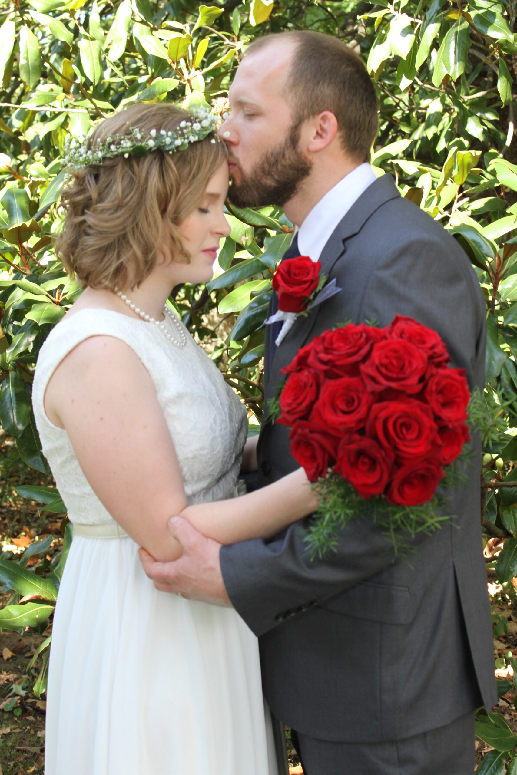Groom, wearing a grey suit and red rose boutonniere, kissed the bride on the forehead. The bride, wearing a white lace sundress and flower crown, holds a large bouquet of red roses. 