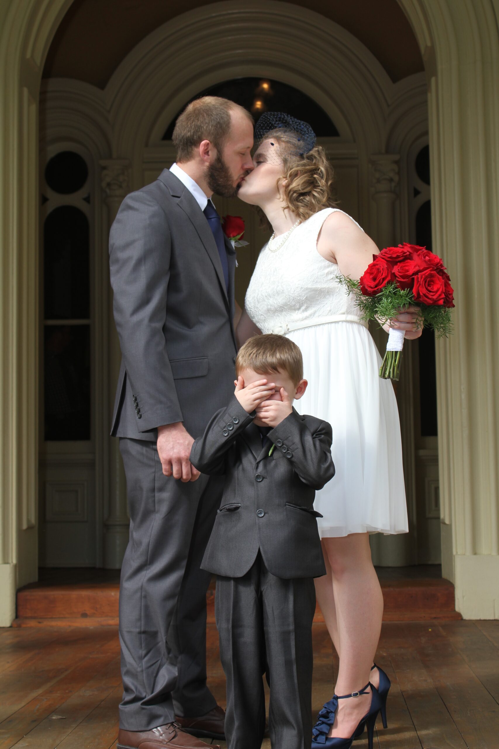 Groom, wearing a grey suit, blue tie and red rose boutonniere kisses the bride. The bride, wearing a white lace sundress and navy bird cage veil with navy ruffled heels, holds a large bouquet of red roses. Their son, wearing a gray suit and navy tie covers his eyes as his parents kiss. 