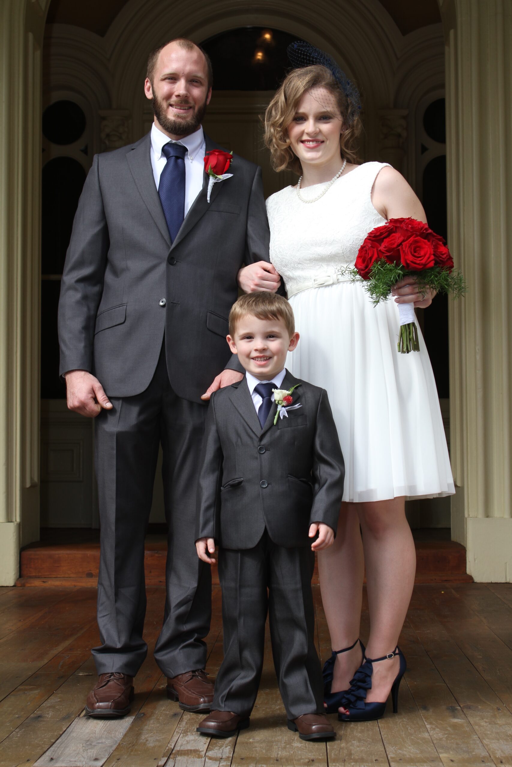 Groom, wearing a grey suit, blue tie and red rose boutonniere links arms with the bride. The bride, wearing a white lace sundress and navy bird cage veil with navy ruffled heels, holds a large bouquet of red roses. Their son, wearing a gray suit and navy tie smiles at the camera. The ring bearer has a small white rose boutonniere.