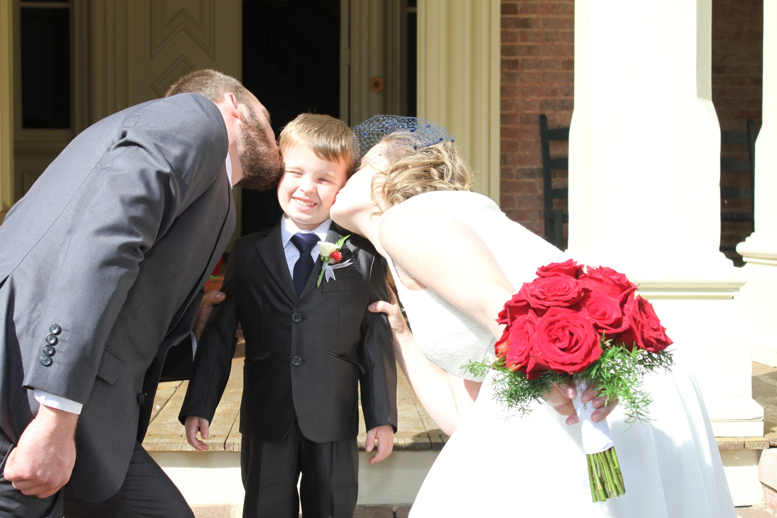 Groom, wearing a grey suit, blue tie and red rose boutonniere and the bride, wearing a white lace sundress and navy bird cage veil with navy ruffled heels, holding a large bouquet of red roses lean down to kiss their son on each cheek. The ring bearer is wearing a gray suit and navy tie smiles at the camera. The ring bearer has a small white rose boutonniere.
