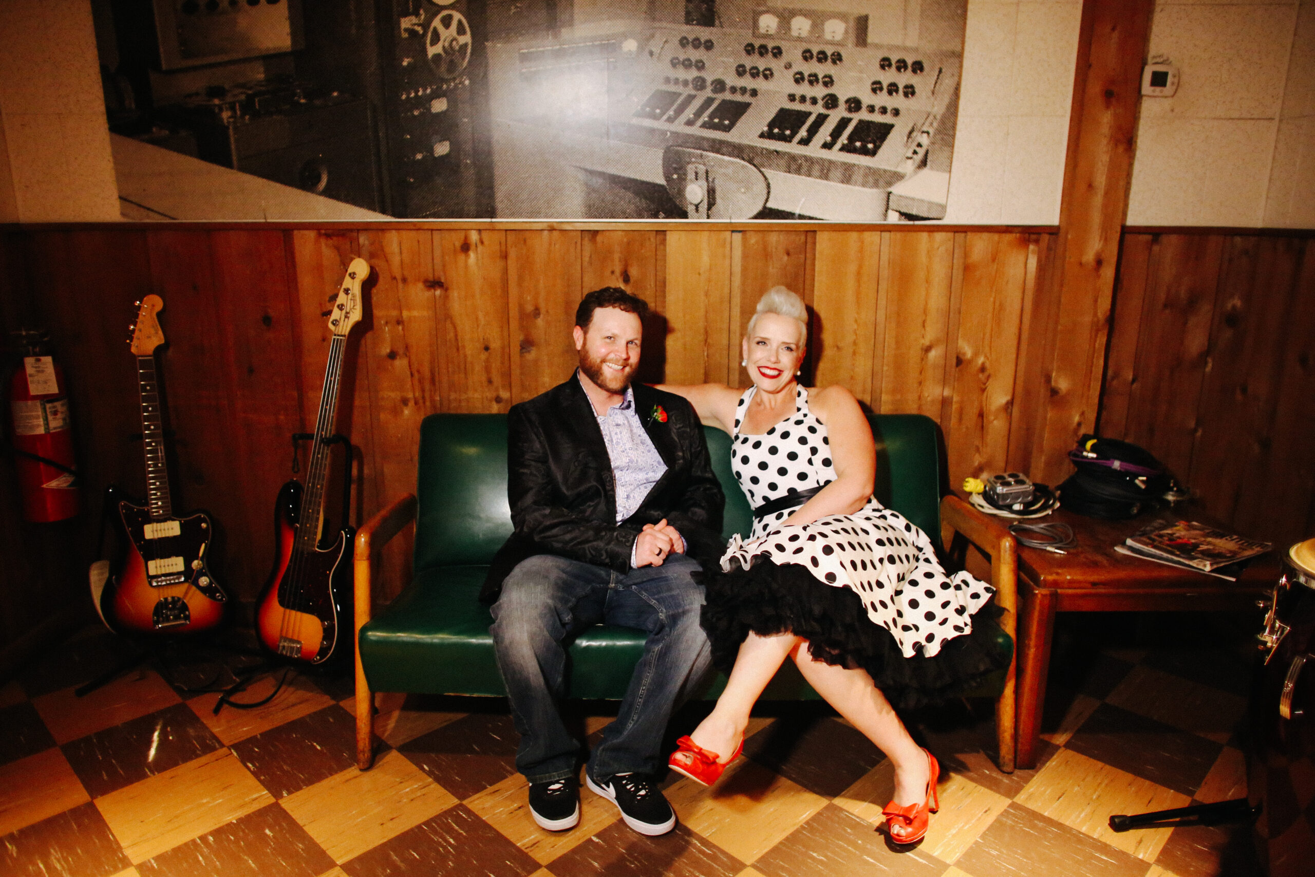 The bride and groom sit on a black mid-century couch with wooden arms at RCA Studio B. The bride has her arm around the back of the couch. The floor is black and white checkered linolium. The walls are half wood paneling and half ivory wallpaper. Above their heads is a large black and white photo of a sound board at RCA Studio B. On the left of the couch are two electric guitars on stands. To the right of the couch is a small wooden end table with a black telephone, two magazines and an electrical outlet. The bride is wearing a halter neck white pinup dress with black polka dots. She has a black belt and a black crinoline petticoat with large pearl earrings. The bride has bright red lipstick to match her bright red peep toe shoes with a small bow. The groom is wearing a black silk suit jacket with a paisley pattern, a white shirt with small blue flowers, a red rose boutonniere and dark washed jeans and black sneakers. 