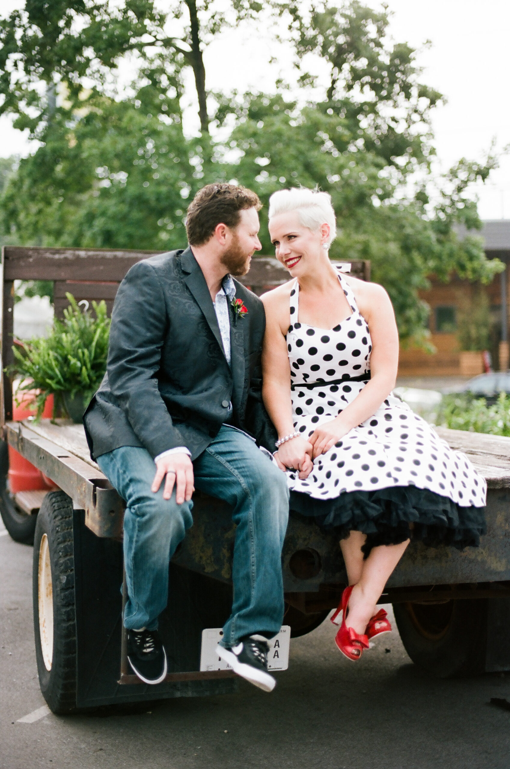 Sitting on the back of a flatbed pickup truck with ferns in the back, the bride and groom gaze into each other's eyes. The bride is wearing a halter neck white pinup dress with black polka dots. She has a black belt and a black crinoline petticoat. The bride has bright red lipstick to match her bright red peep toe shoes with a small bow. The groom is wearing a black silk suit jacket with a paisley pattern, a white shirt with small blue flowers, a red rose boutonniere and dark washed jeans. 