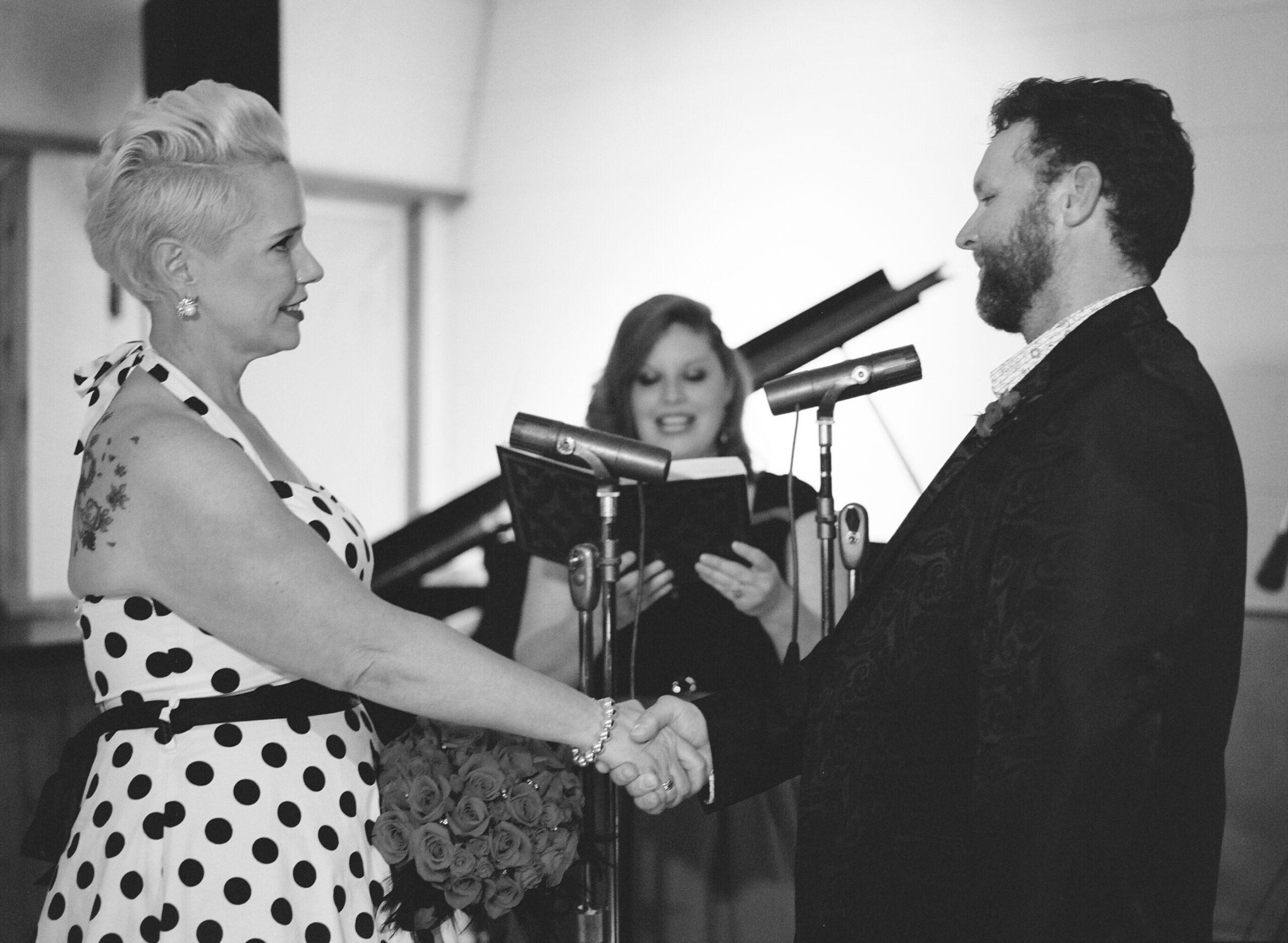 A black and white image of the wedding ceremony. With the officiant behind them holding a leather bound book, the bride and groom speak into microphones as they say their vows while holding hands. The groom and bride look into each other's eyes. The bride is wearing a halter neck white pinup dress with black polka dots. She has a black belt and a black crinoline petticoat. Her rose bouquet is accented with rhinestones and black feathers. She has pearl and rhinestone earrings and a silver beaded bracelet. The groom is wearing a black silk suit jacket with a paisley pattern, a white shirt with small flowers, a boutonniere. 