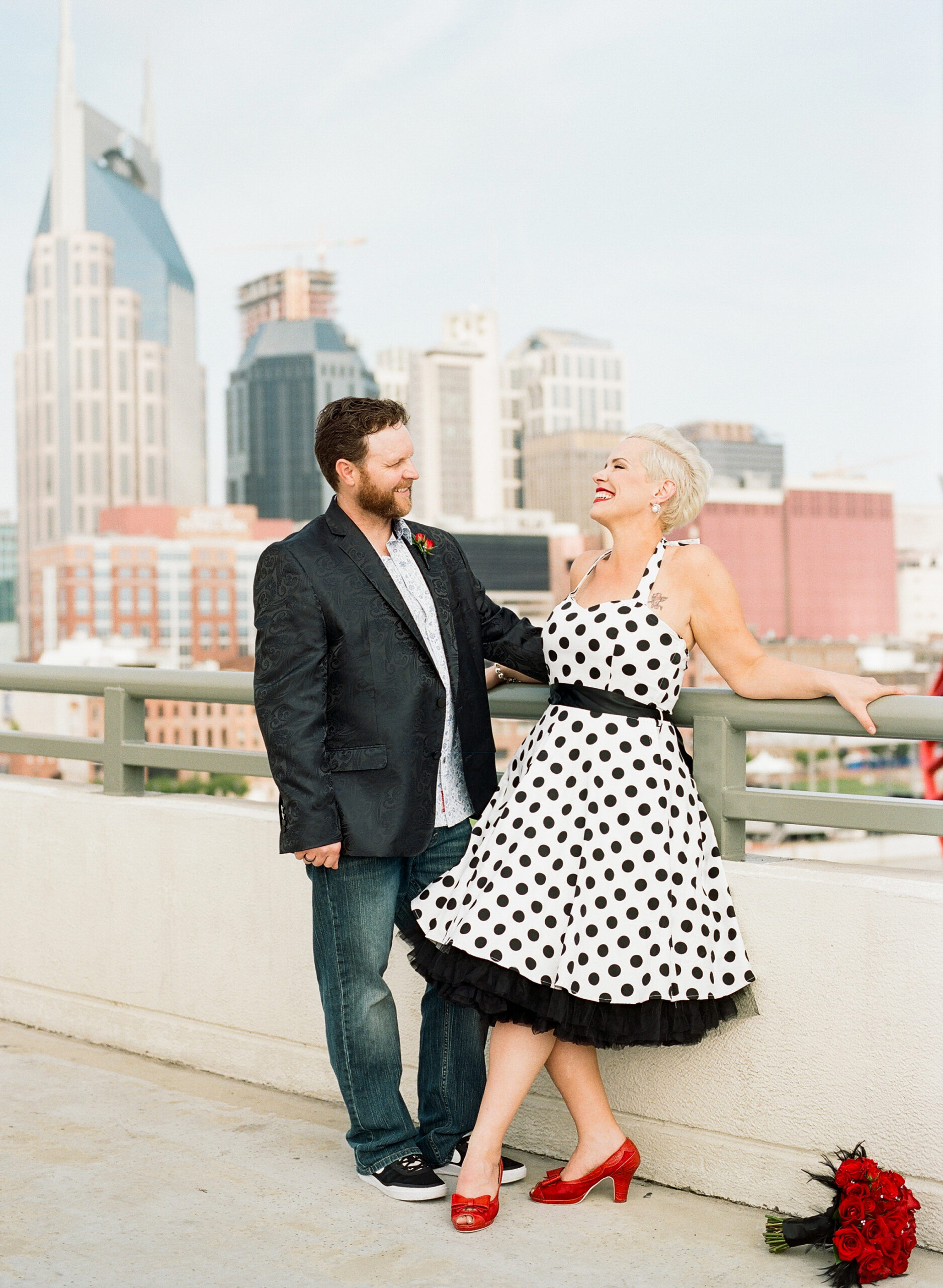 Standing on the pedestrian bridge with the Nashville skyline in the background, The bride laughs at the groom as she leans back on the railing. She is wearing a halter neck white pinup dress with black polka dots. She has a black belt and a black crinoline petticoat. The bride has bright red lipstick to match her bright red peep toe shoes with a small bow. Her bright red rose bouquet accented with rhinestones and black feathers is on the ground by her feet. The groom is wearing a black silk suit jacket with a paisley pattern, a white shirt with small blue flowers, a red rose boutonniere and dark washed jeans. 
