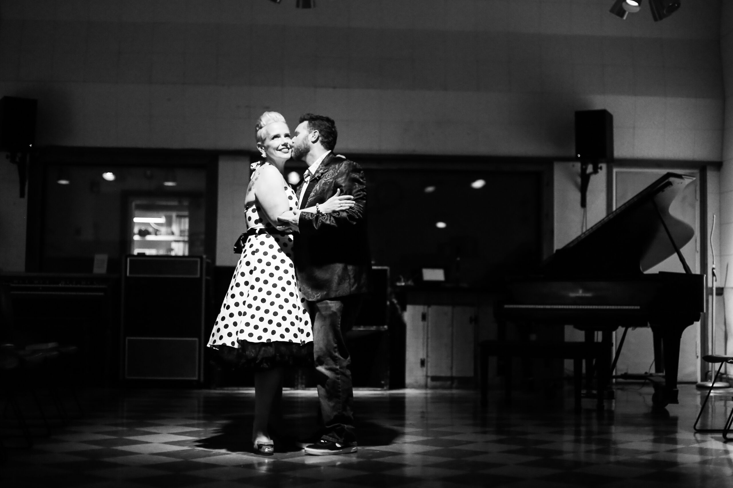 A black and white image of the bride and groom dancing under a spotlight on a black and white checkered floor. An opened grand piano is in the back ground on the right. The window to the audio engineer's room is lit up on the left. The bride is wearing a halter neck white pinup dress with black polka dots. She has a black belt and a black crinoline petticoat. The bride has peep toe shoes with a small bow. She has pearl and rhinestone earrings and a silver beaded bracelet. The groom is wearing a black silk suit jacket with a paisley pattern, a white shirt with small blue flowers, a red rose boutonniere with dark washed jeans and black sneakers. She is smiling while looking upwards as he leans in to kiss her cheek. 