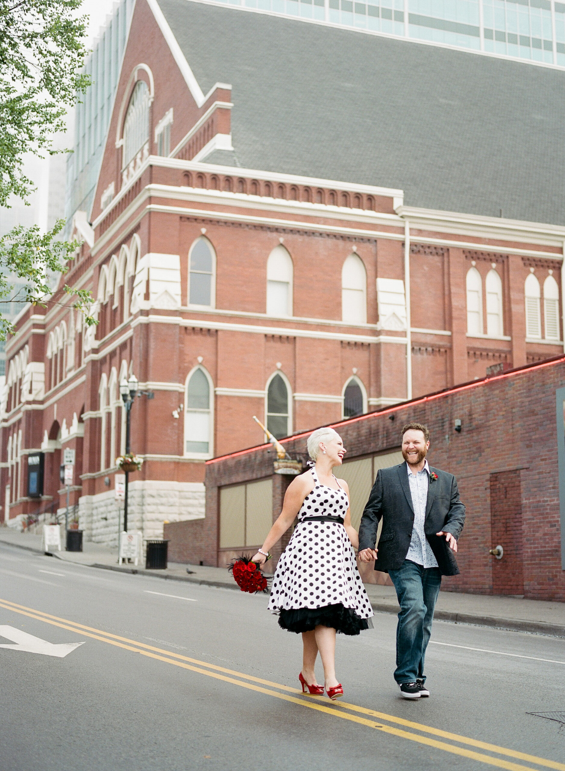 Walking down 2nd Avenue in Nashville along side the Ryman Auditorium, the bride and groom hold hands laughing as they look at each other. The bride is wearing a halter neck white pinup dress with black polka dots. She has a black belt and a black crinoline petticoat. The bride has bright red lipstick to match her bright red peep toe shoes with a small bow and her bright red rose bouquet accented with rhinestones and black feathers. The groom is wearing a black silk suit jacket with a paisley pattern, a white shirt with small blue flowers, a red rose boutonniere and dark washed jeans. 