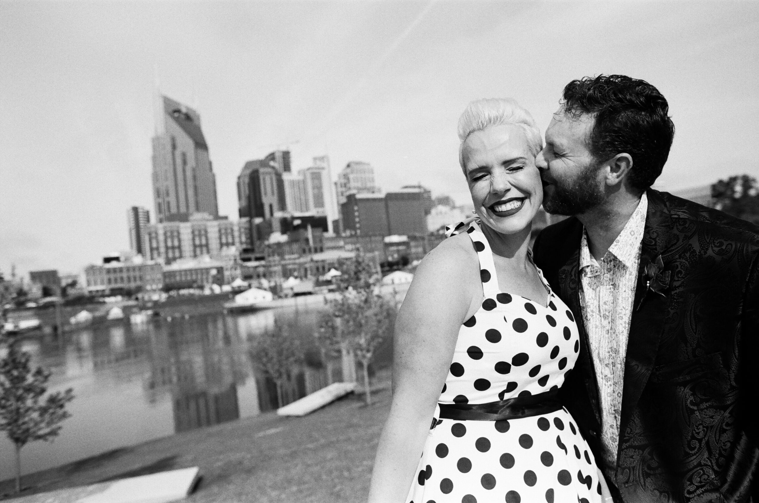 A black and white image of the groom kissing the bride's cheek and the bride closes her eyes and smiles big. The Cumberland River flows behind them with the Nashville skyline in the background. The bride is wearing a halter neck white pinup dress with black polka dots and a black belt. The groom is wearing a black silk suit jacket with a paisley pattern, a white shirt with small flowers. 
