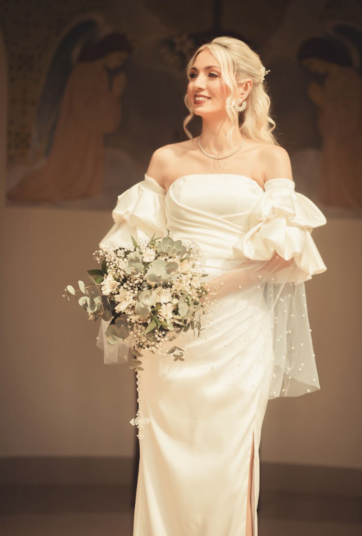 Blonde bride in an satin wedding dress with elaborately ruffled sleeves with pearl beaded tulle and an off the shoulder bodice. 