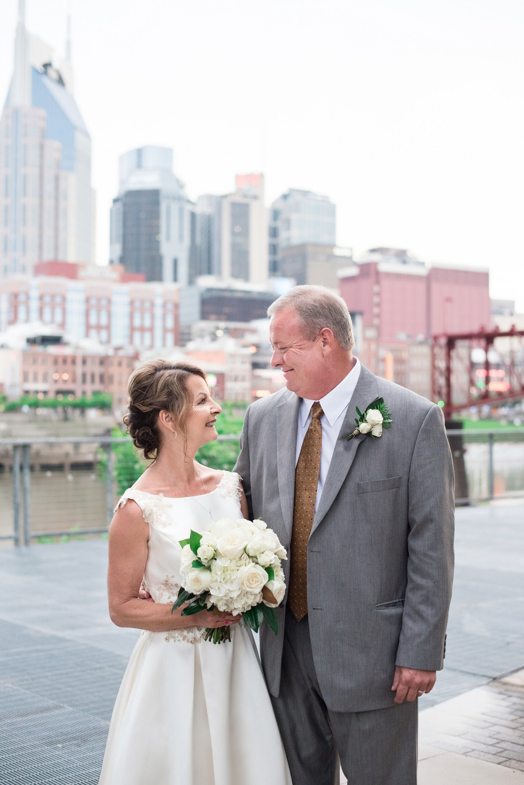 The bride, wearing a tea length white wedding dress with lacy floral accents holds a large white bouquet of roses and hydrangea. She is smiling up at the groom who is wearing a gray suit with a white shirt and brown tie. He has a white boutonniere pinned to his lapel. He is smiling down at the bride. The Cumberland River and Nashville skyline are in the background behind them. 