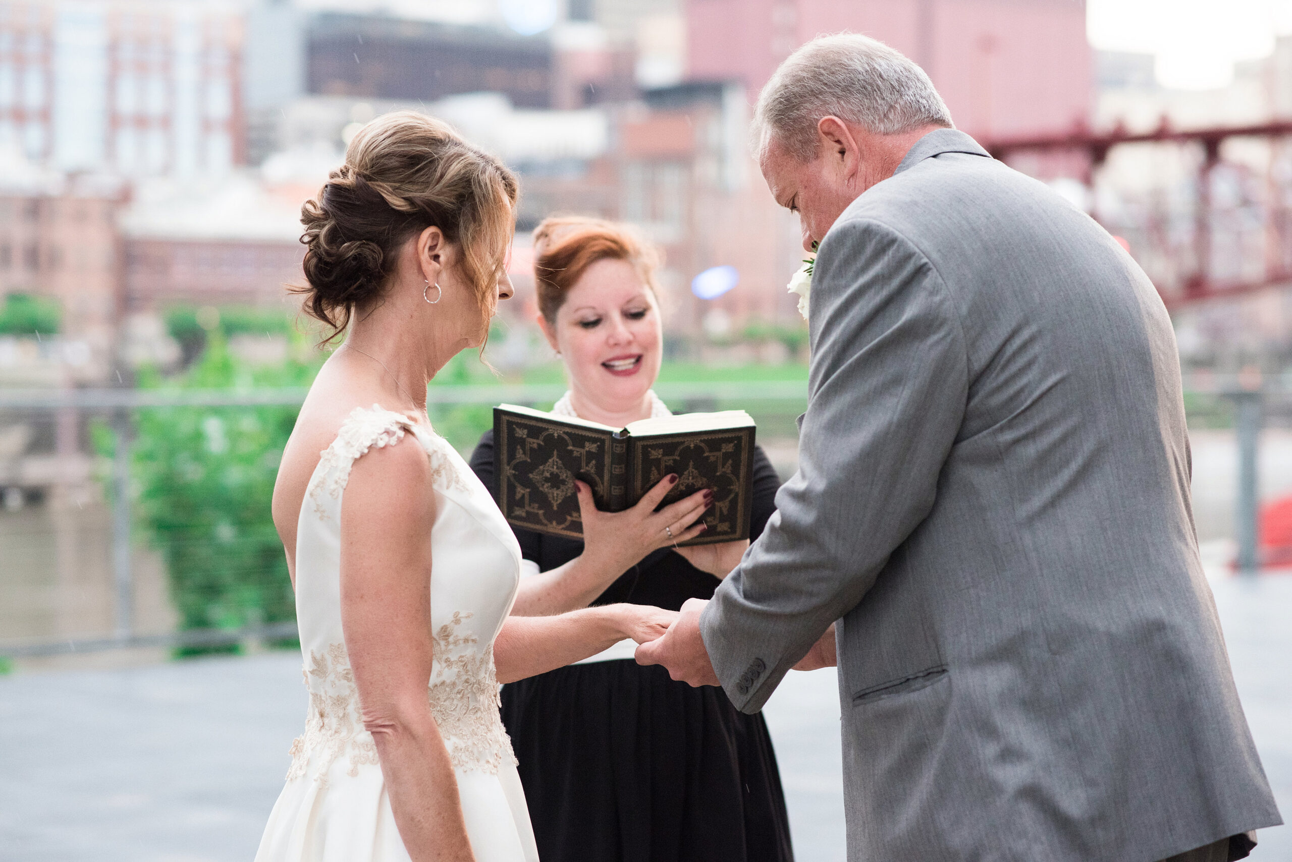 The officiant stands before the bride and groom wearing a black dress holding a leather bound book with gold detail. The bride holds out her hand as the groom places the wedding ring on her finger. The bride is wearing a white sleeveless wedding dress with a plunging back and lace detailing around the waist and shoulders. The groom is wearing a gray suit with a white boutonniere. 