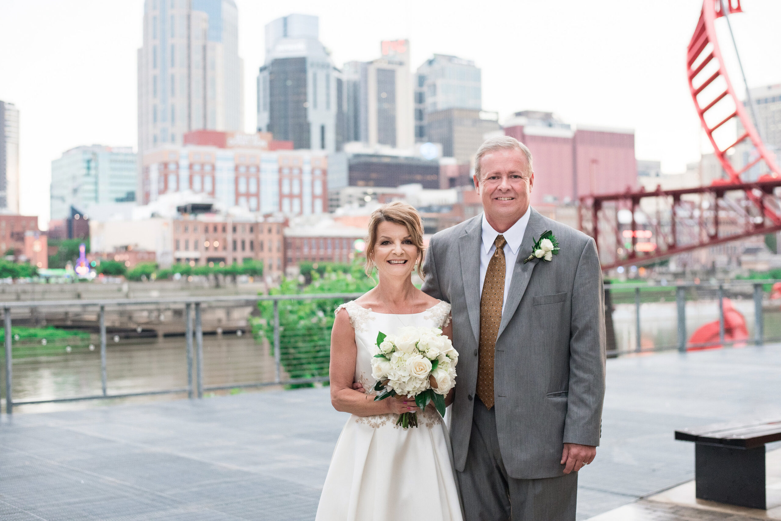 The bride and groom smile at the camera with the skyline and Cumberland River behind them. The bride is wearing a tea length white sleeveless wedding dress with lacy floral accents. The groom  is wearing a gray suit with a white shirt and brown tie and a white boutonniere. The bride is holding a large white bouquet of roses and hydrangea. 