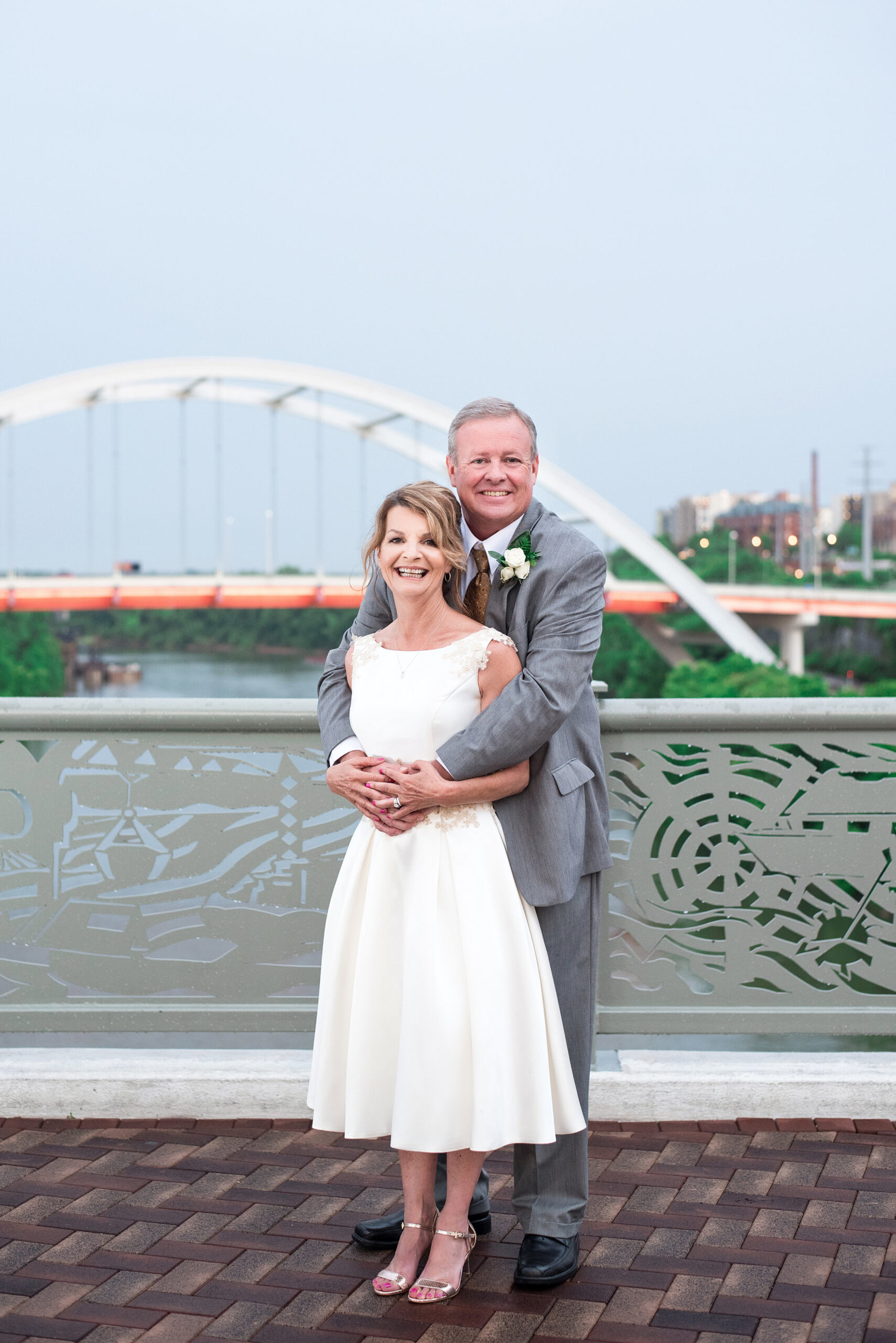 Standing on the Pedestrian Bridge in Nashville with another bridge in the background, the groom embraces the bride from behind as they both smile for the camera. The bride is wearing a tea length white sleeveless wedding dress with lacy floral accents and gold stiletto heels. The groom  is wearing a gray suit with a white shirt and brown tie and a white boutonniere. The Cumberland River and Nashville skyline are in the background behind them. 