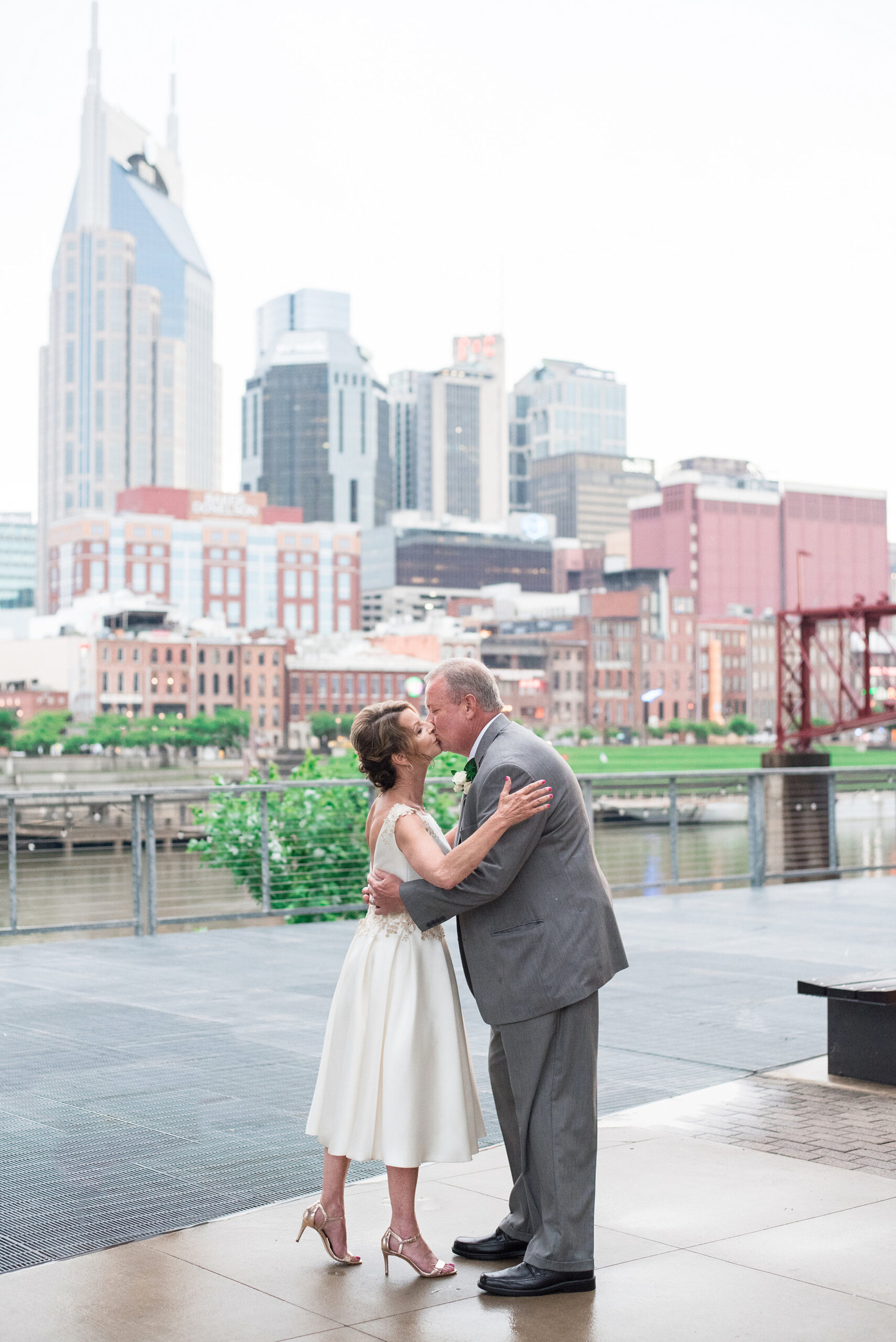 The bride, wearing a tea length white wedding dress with lacy floral accents and strappy gold stillettos embraces and kisses the groom who is wearing a gray suit with a white shirt. He has a white boutonniere pinned to his lapel. The Cumberland River and Nashville skyline are in the background behind them. 