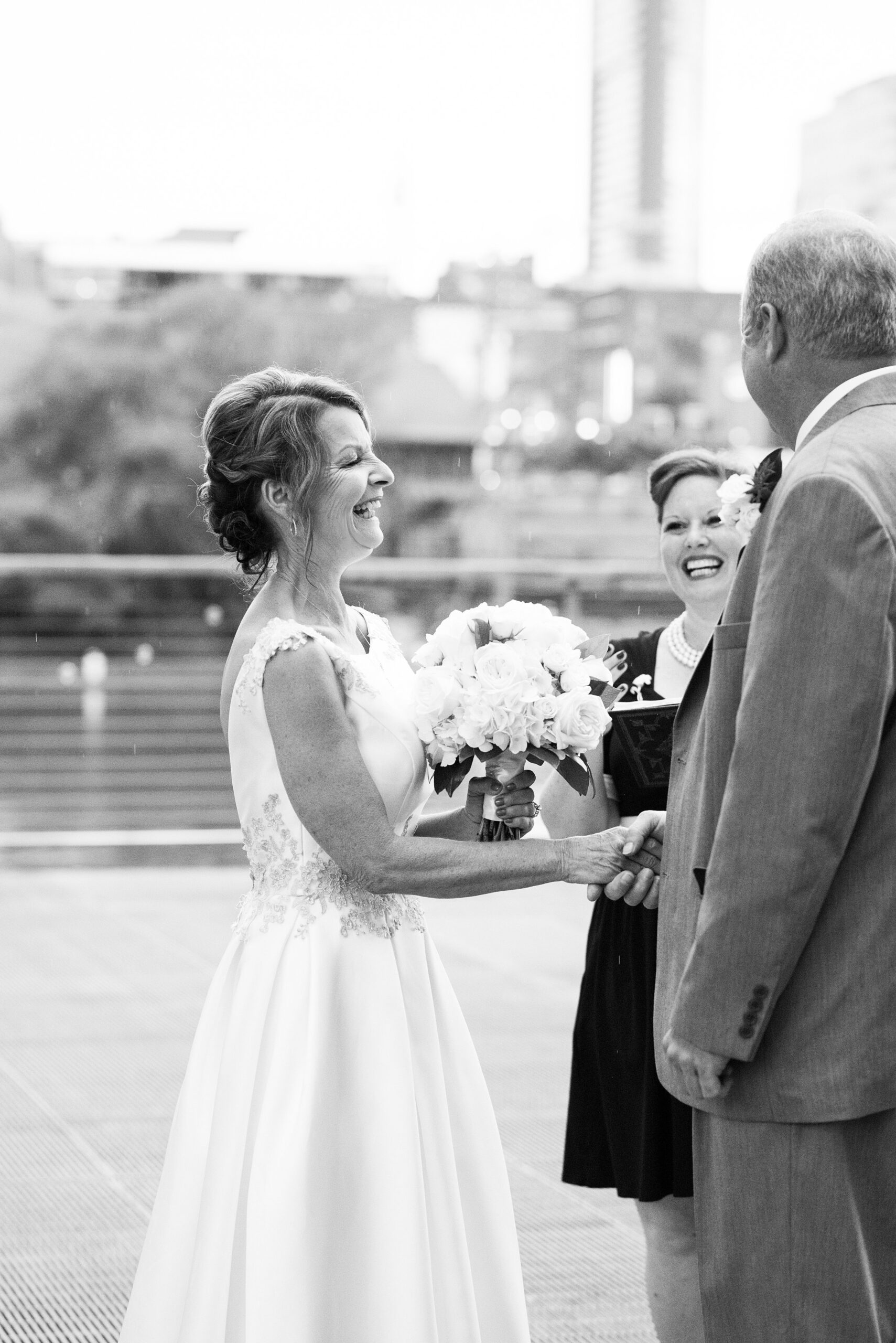 A black and white image of the bride, groom and officiant laughing during the ceremony. The bride and groom hold hands and the bride holds her large bouquet of roses, hydrangeas and magnolia leaves. The bride is wearing a sleeveless wedding dress with a plunging back and lace detailing on the shoulders and around the waist. The groom is wearing a suit with a white shirt and white boutonniere. The officiant is wearing a black dress with three strands of pearls. 