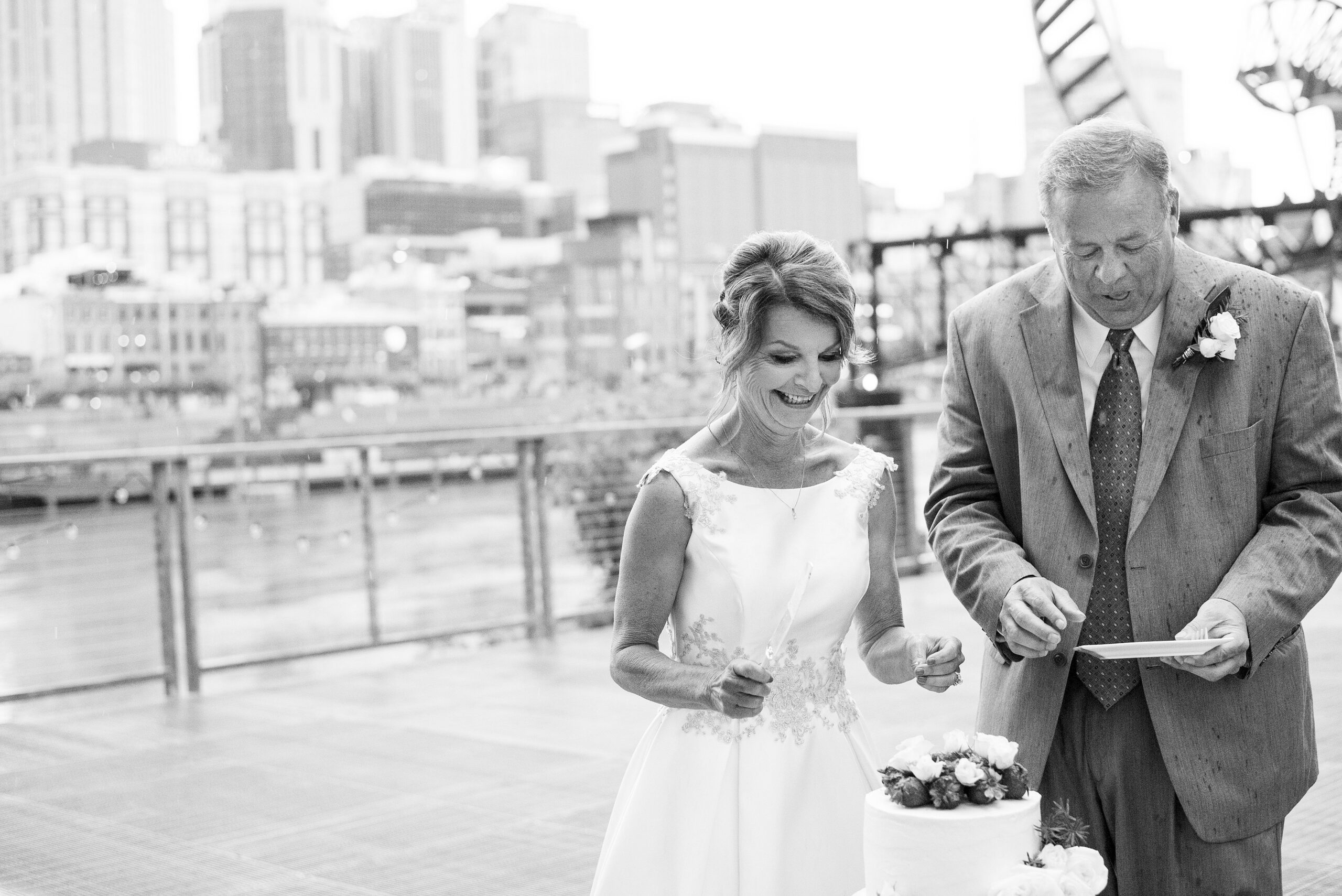 A black and white photo of the bride smiling down at the cake while holding a cake knife. The groom holds out a white square plate for her to put the cake on. The cake is a small one tier cake with strawberries and white flowers. The bride is wearing a tea white sleeveless wedding dress with lace detailing around the waist. The groom is wearing a suit with a white shirt and tie. The groom has a white boutonniere. The Cumberland River and Nashville skyline are in the background behind them.