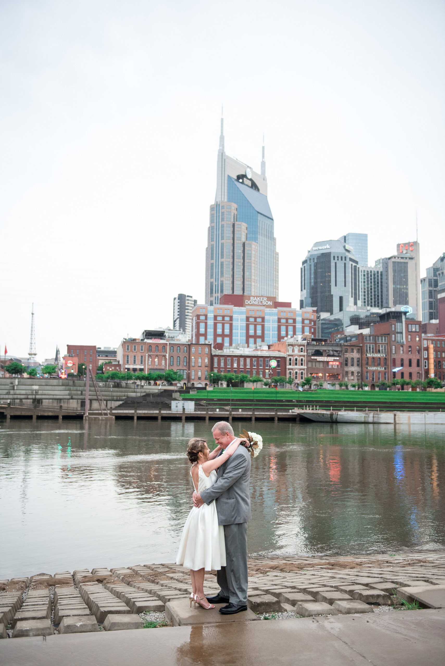 Standing at the riverfront smiling up at each other the bride and groom embrace. The bride is wearing a tea length sleeveless wedding dress with a plunging back and gold strappy heels. The groom  is wearing a gray suit with black shoes. The Cumberland River, the Batman Building and Nashville skyline are in the background behind them. The bride has her arms around the groom's neck and is holding a large white bouquet of roses and hydrangea with magnolia leaves.
