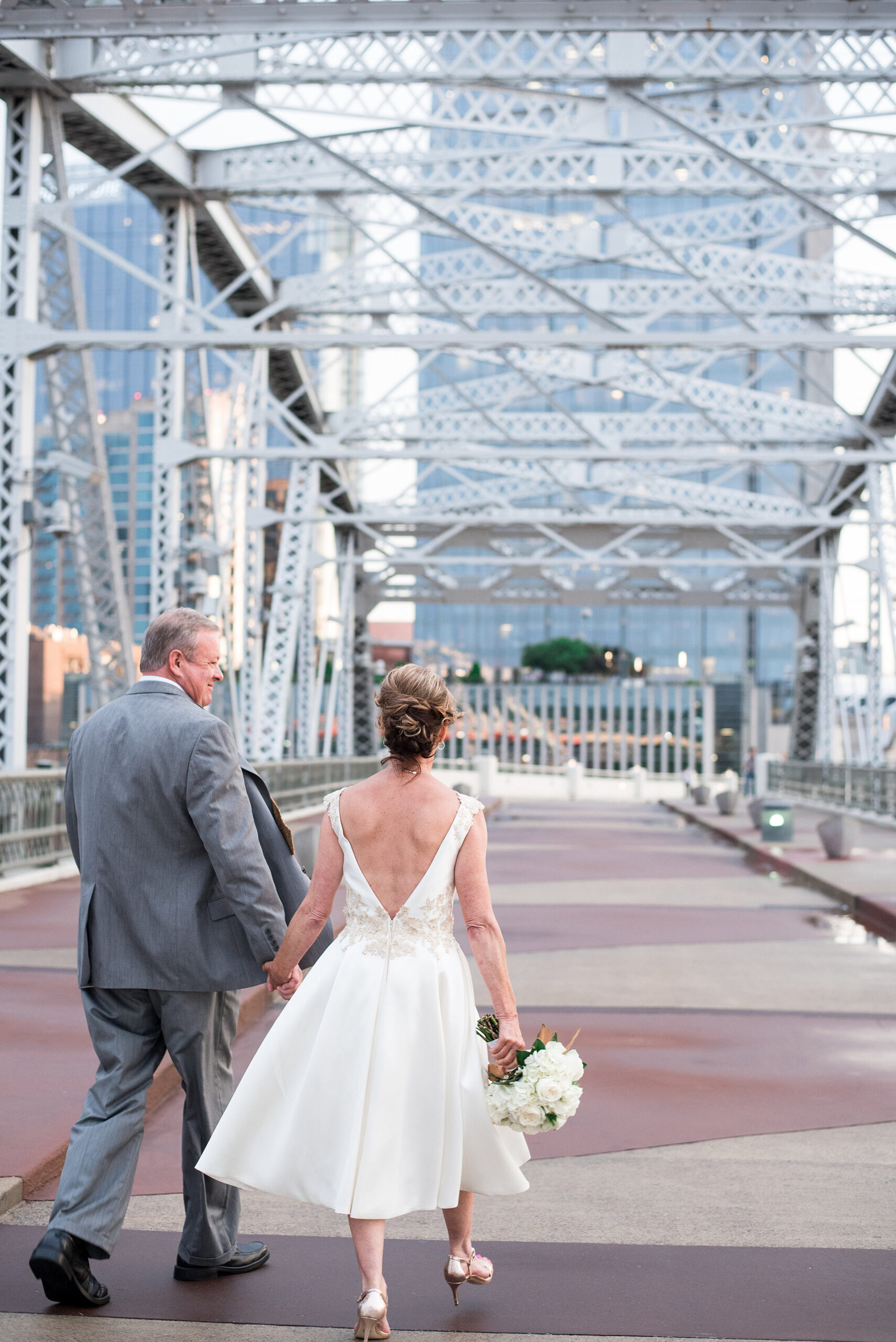 Walking hand in hand across the Pedestrian Bridge in Nashville, the bride and groom smile at each other. floor is red and beige striped in a triangle pattern. The grey trusses of the bridge are above them. The bride is wearing a tea length white wedding dress with a plunging back and no sleeves. There is lace detailing around the waist and at the shoulders. The bride has gold stilettos and is holding a large white bouquet of hydrangea and roses with magnolia leaves. The groom is wearing a gray suit with a white shirt and black shoes. 