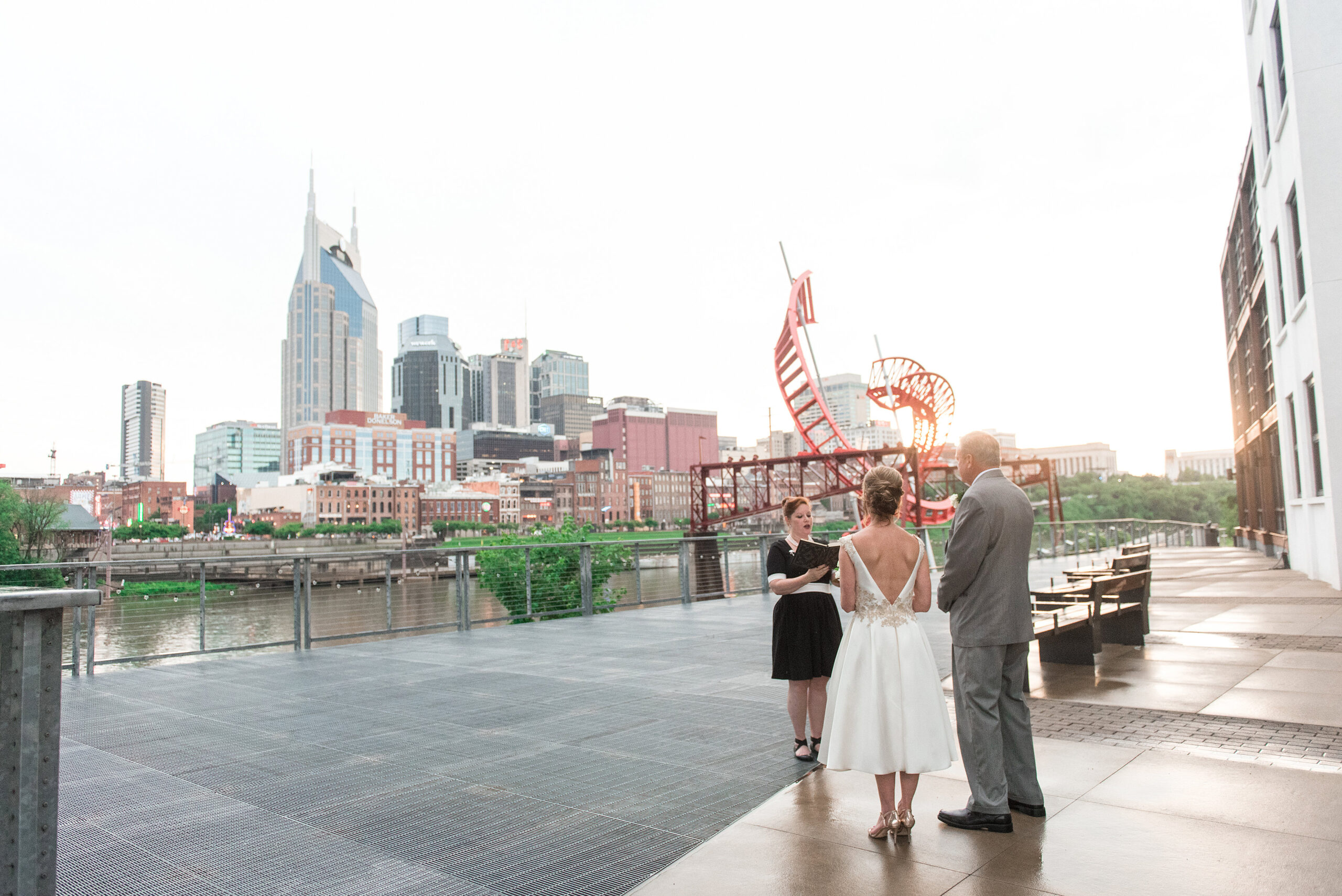 With the Nashville skyline behind them, the bride and groom listen to the officiant during their wedding ceremony. Standing on the banks of the Cumberland River the officiant is wearing a black dress with a white belt and black shoes. She is holding a leather bound book. The bride is wearing a tea length white sleeveless wedding dress with a plunging back and lace detailing around the waist and shoulders. The groom is wearing a gray suit with black shoes. 