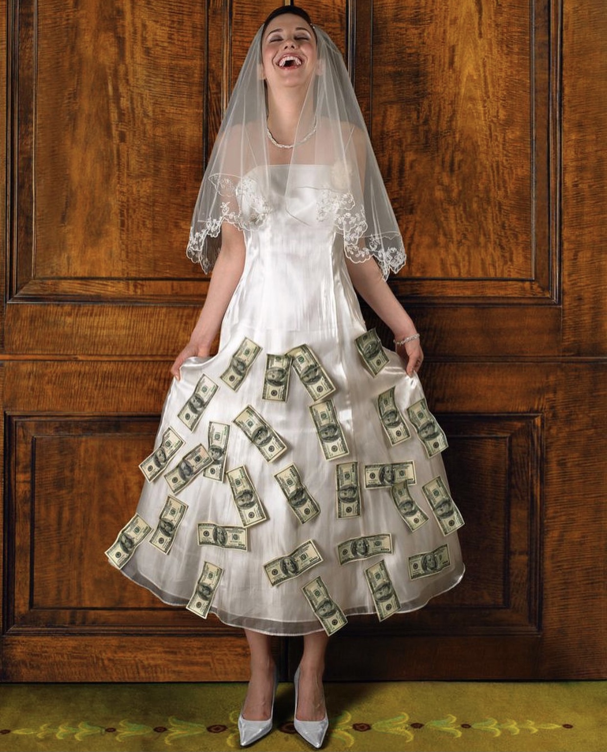 Bride wearing a satin ankle length wedding dress with an organza overlay and an elbow length veil trimmed in lace tilts her face to the sky laughing. She has $100 dollar pills pinned all over the skirt of her dress. She is standing in front of a large wood door. 