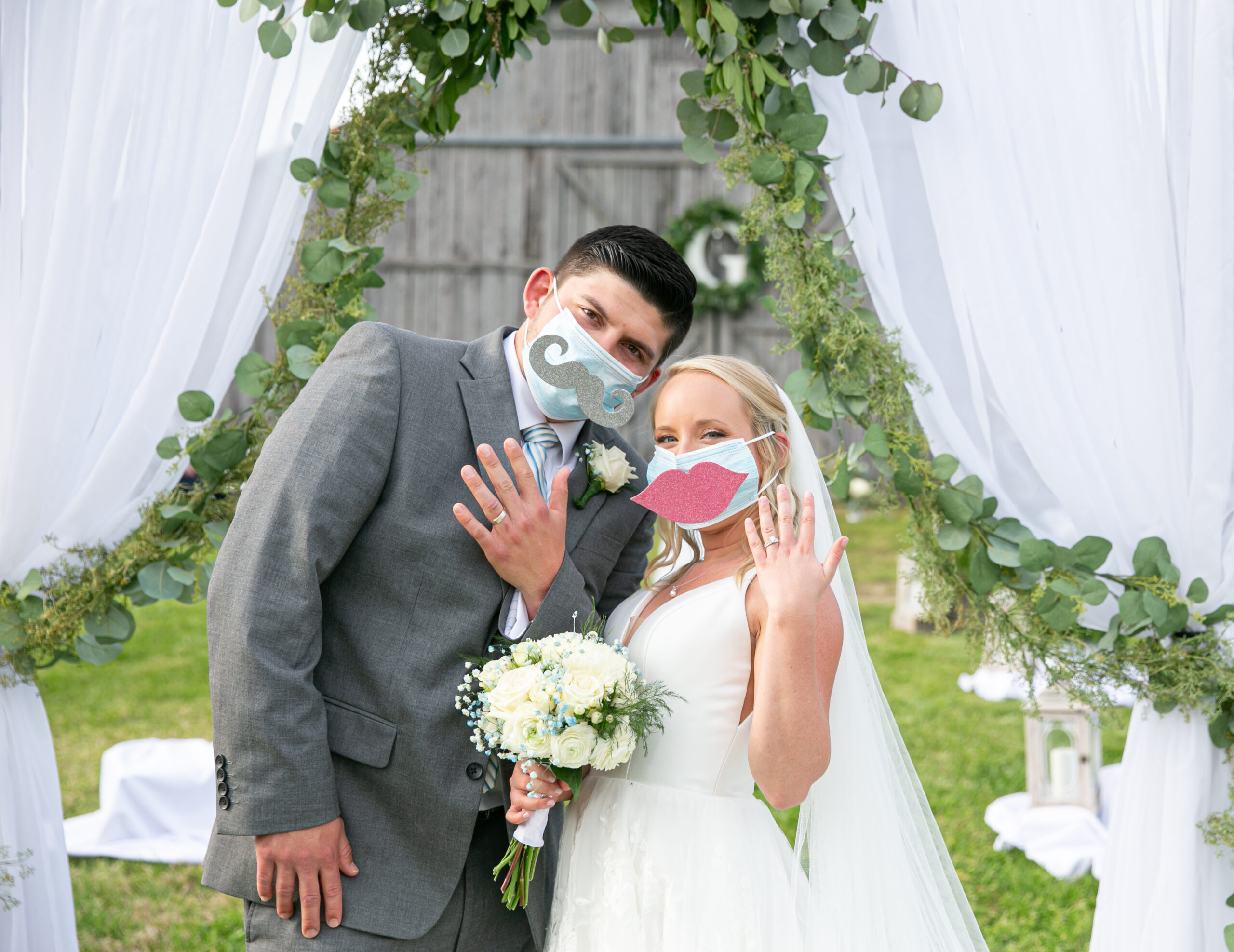 COVID bride and groom poses underneath an arbor of white drapery decorated with a green garland. The bride and groom show off their wedding rings. The groom is wearing a gray suit with a white shirt and light blue and white striped tie with a light blue medical mask that has a large gray felt mustache attached to the front. The bride is wearing a light blue medical mask that has a large pink felt set of lips attached to the front. The bride is holding a bouquet of white and blue flowers. 