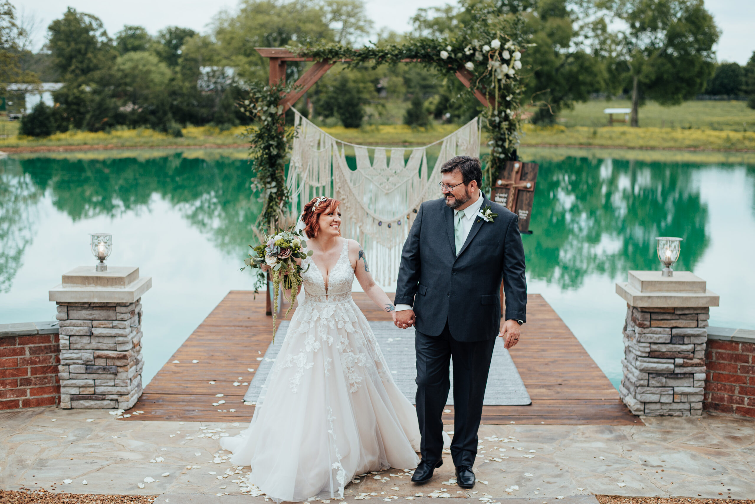Bride wearing a flowing wedding dress with lace accents and a plunging neckline holds hands with the groom who is wearing a navy suit and light green tie. They are walking away from the pier over the pond at Steel Magnolia Barn. their arbor is decorated with a large macreme piece, greenery and white flowers. There are stone pillars and low brick walls on either side of them. 