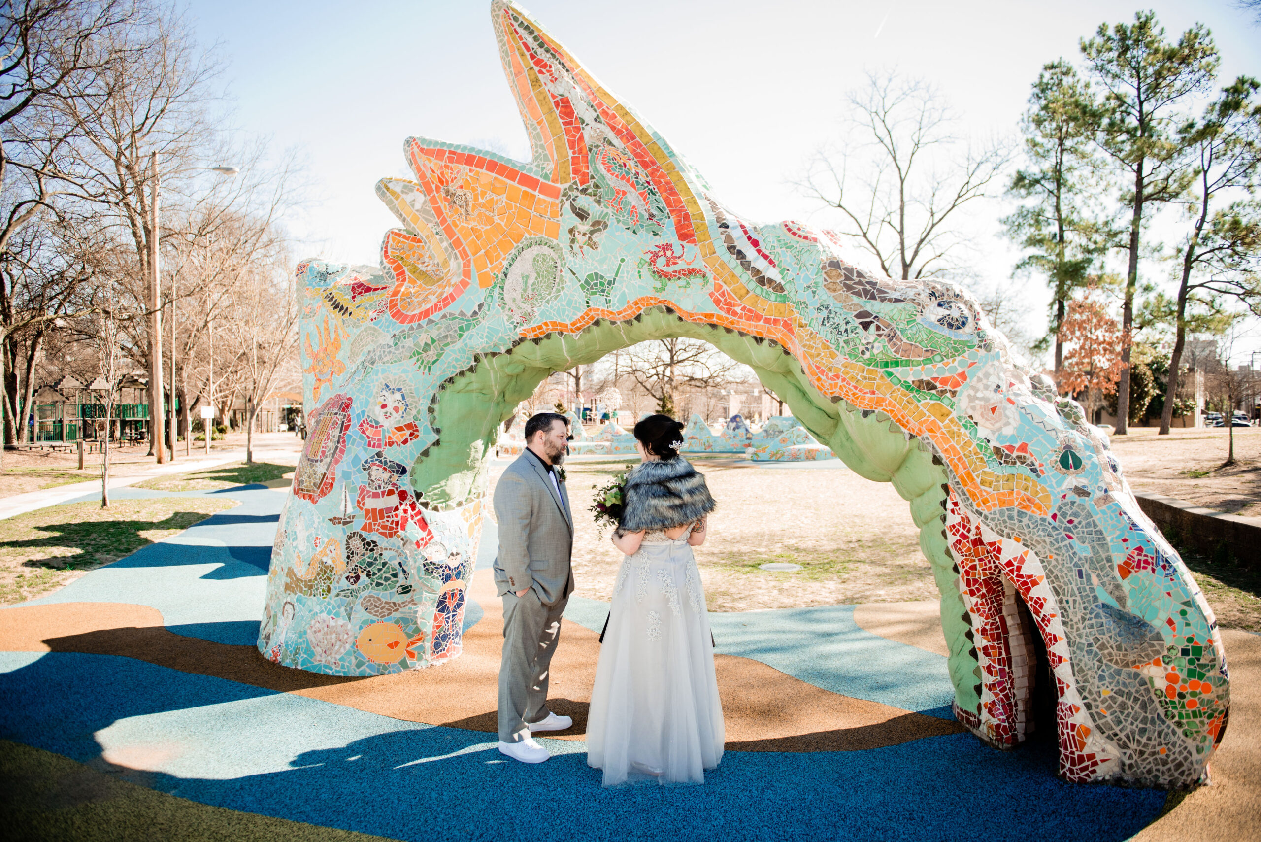 The bride, wearing a lace silver dress, and gray fur wrap holds a large bouquet as she faces the groom. The groom, wearing a gray suit with a black shirt, white tie and white and mauve boutonniere stands with his hands in his pockets facing the bride. They stand beneath a large mosaic dragon at Dragon Park in Nashville. 