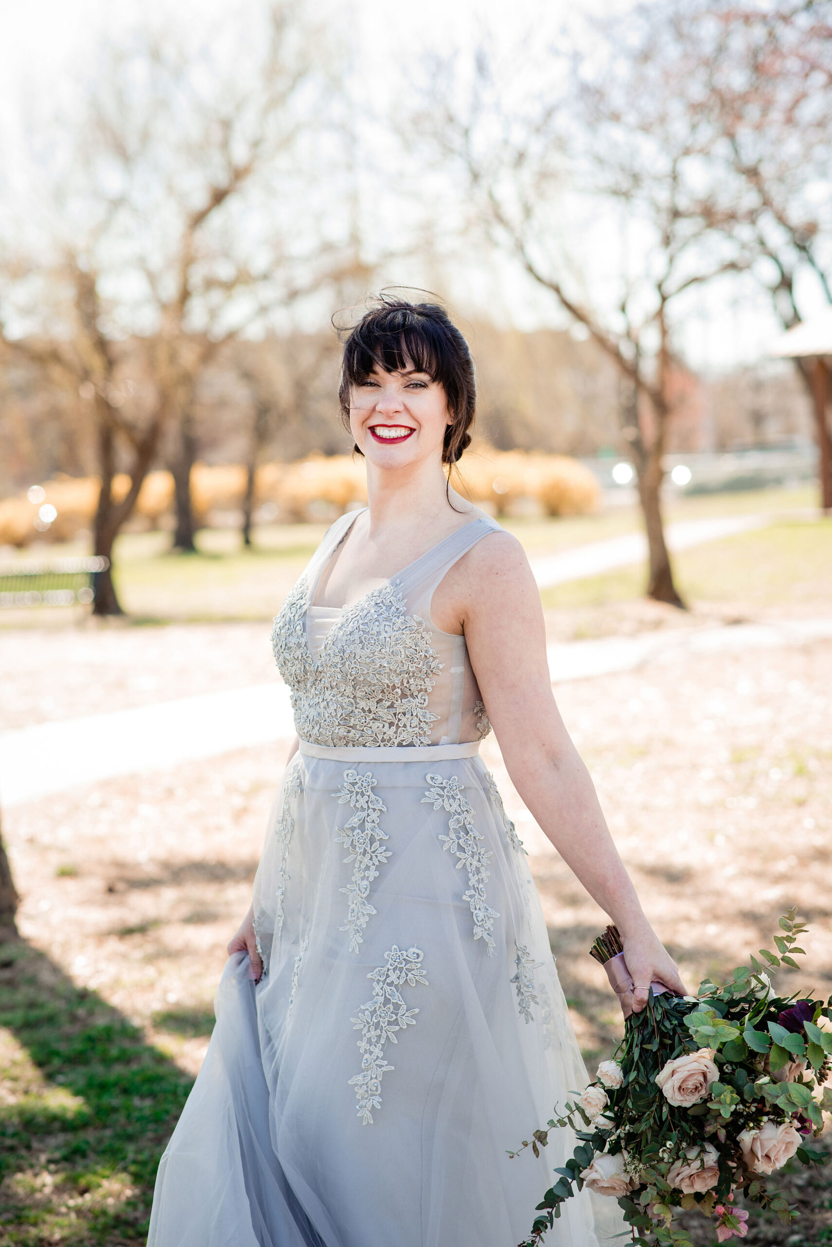 The bride is wearing a v-neck silver lace wedding dress with a tulle skirt and floral appliques. She is holding a large bouquet of eucalyptus with blush roses and pink berries. 