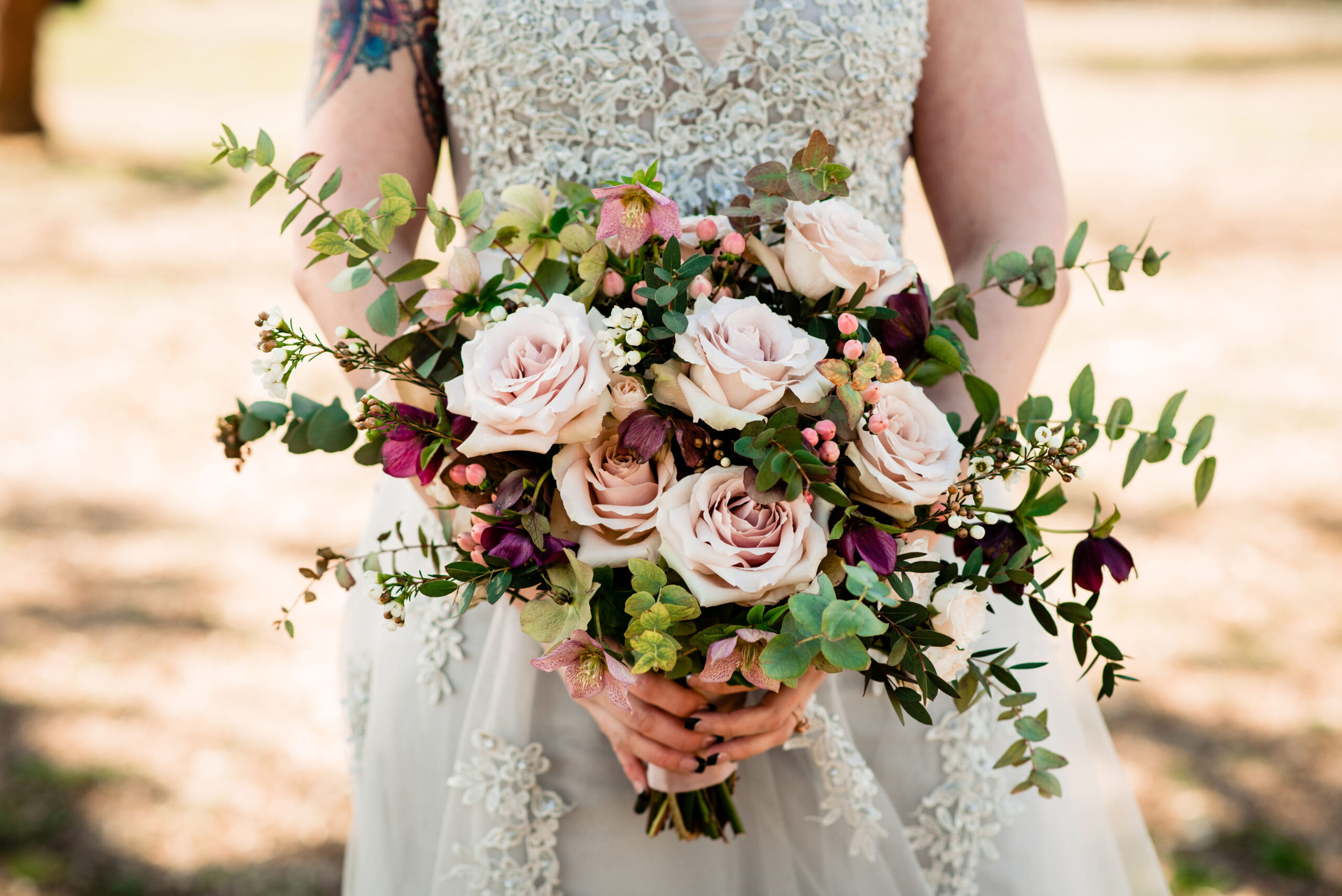 The bride, wearing a v-neck silver lace wedding dress with a tulle skirt and floral appliques. She holds a large bouquet of eucalyptus, blush roses, magenta and pink flowers and pink berries. 