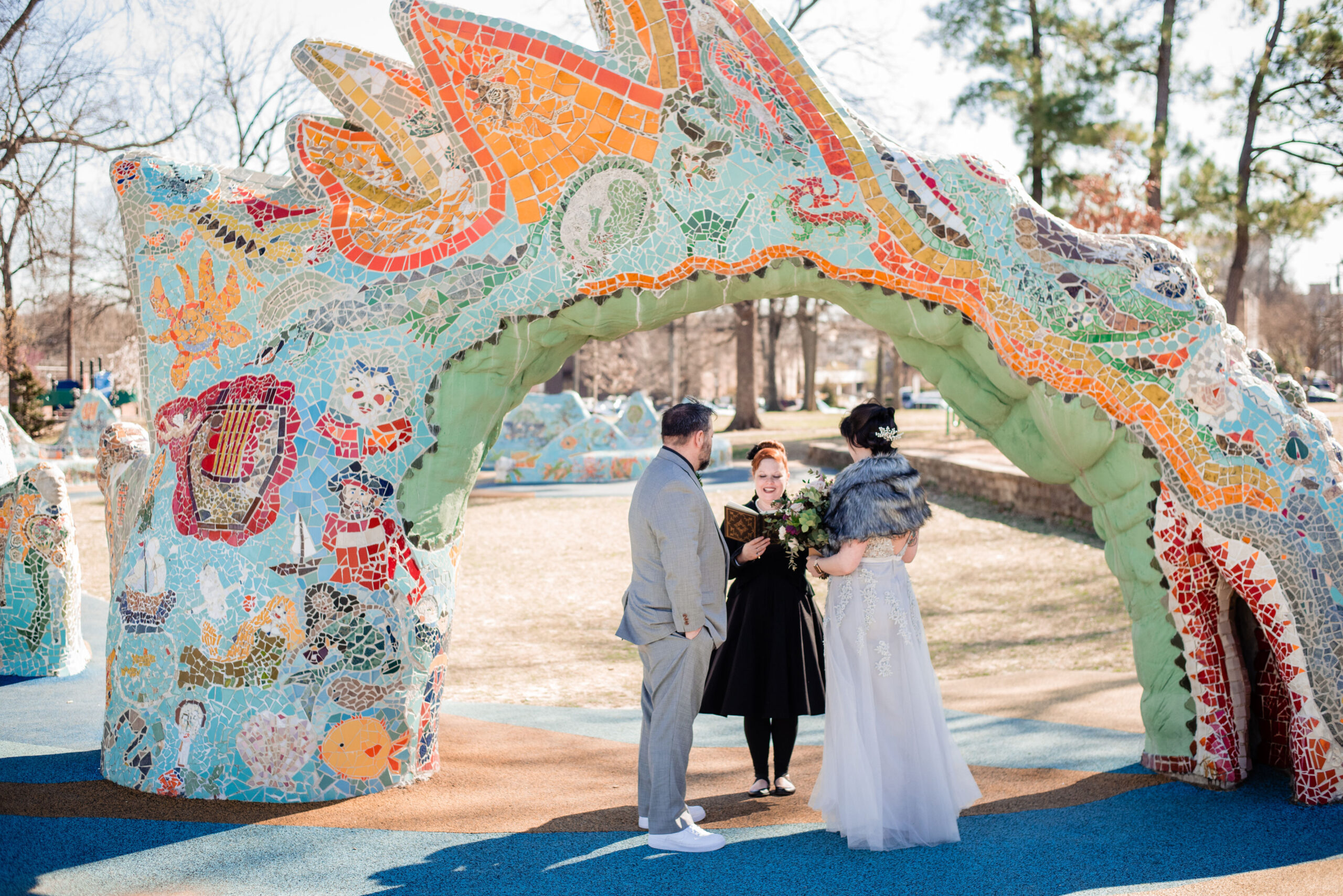 The bride, wearing a lace silver dress, and gray fur wrap holds a large bouquet as she faces the officiant. The groom, wearing a gray suit with a black shirt, white tie and white and mauve boutonniere stands with his hands in his pockets facing the officiant. They stand beneath a large mosaic dragon at Dragon Park in Nashville. The officiant, standing beneath the dragon arbor is wearing a black dress with a black flower in her hair. She holds a leather bound book with gold detailing on the front. 