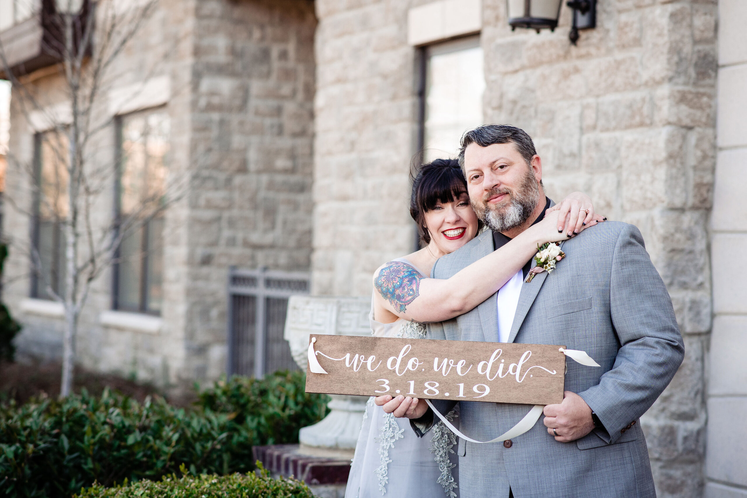 The bride, wearing a lace silver dress, loops her arms around the groom's neck. The groom, wearing a gray suit with a black shirt, white tie and white and mauve boutonniere, holds up a wooden sign announcing their elopement. The sign says, We Do, We Did. They are standing in front of a stone building. 