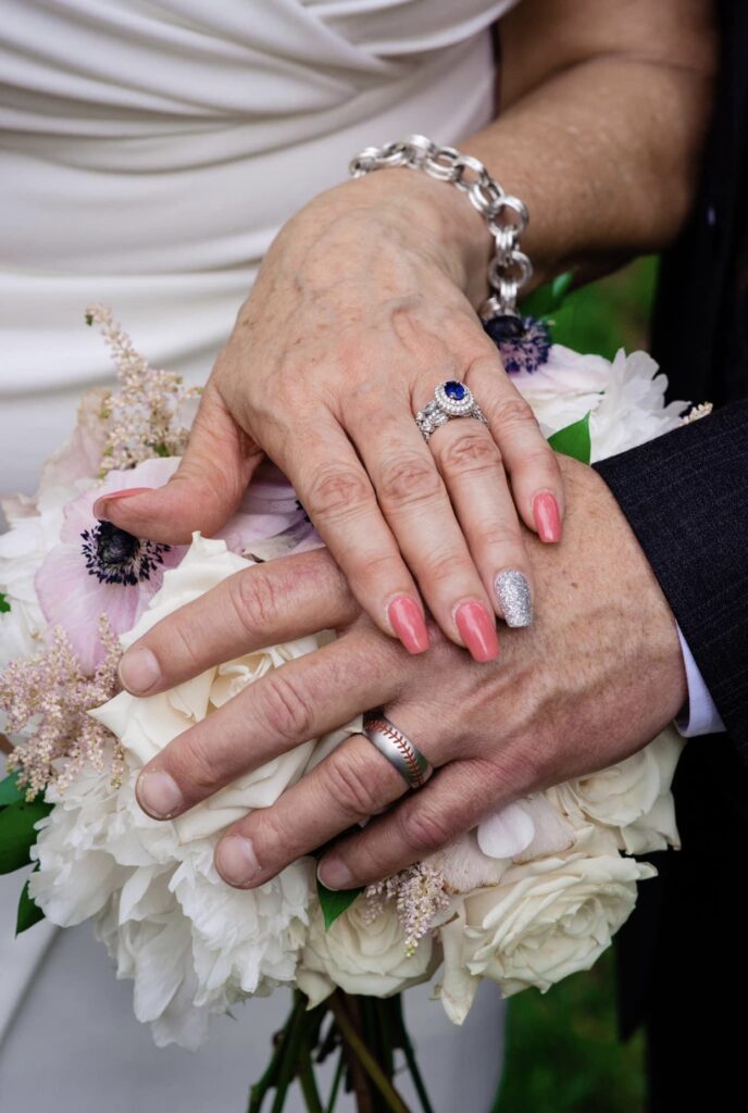 The bride and groom show off their wedding bands. The bride is wearing a fitted white dress with a plunging neckline. Her nails are painted coral and silver. The bride is wearing a silver link bracelet, a diamond wedding band and a sapphire engagement ring. The groom has a baseball themed wedding band. They are holding the bride's bouquet of blush and white flowers. 