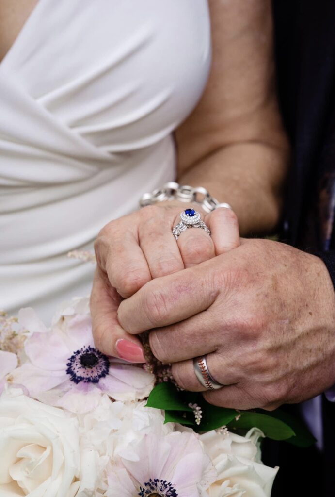 The bride and groom hold hands as they show off their wedding bands. The bride is wearing a fitted white dress with a plunging neckline. Her nails are painted coral and silver. The bride is wearing a silver link bracelet, a diamond wedding band and a sapphire engagement ring. The groom has a baseball themed wedding band. They are holding the bride's bouquet of blush and white flowers. 
