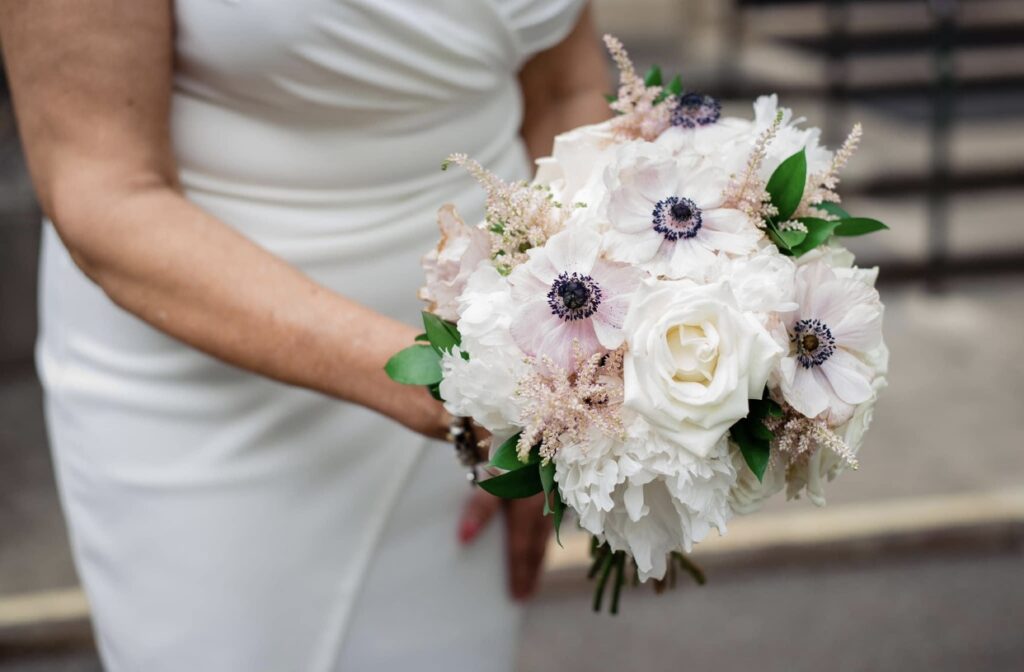 The bride shows off her white, ivory and blush bouquet of peonies, roses and anemonies. The bride is wearing a fitted white dress. 
