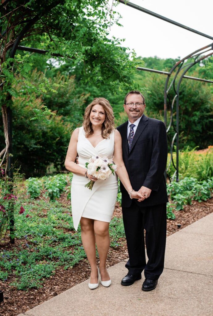 The bride and groom hold hands as they smile at the camera as they stand under an arbor of greenery with orange and yellow flowers in the background. The bride is wearing a fitted white dress with an asymmetrical hemline and plunging neckline. The groom is wearing a black suit with a white shirt and blue tie. The bride is holding a bouquet of white, ivory and blush flowers. They are at Cheekwood Botanical Garden in Nashville.