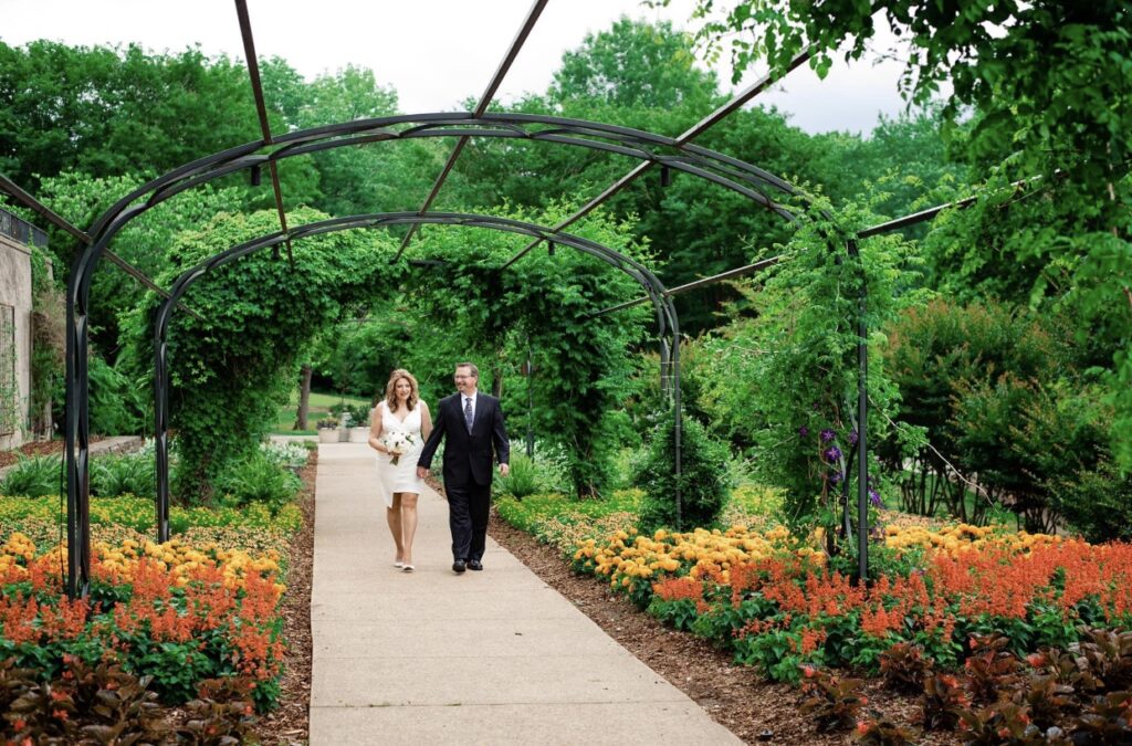 The bride and groom take a stroll under an arbor of greenery with orange and yellow flowers in the background. The bride is wearing a fitted white dress with an asymmetrical hemline and plunging neckline. The groom is wearing a black suit with a white shirt and blue tie. The bride is holding a bouquet of white, ivory and blush flowers. They are at Cheekwood Botanical Garden in Nashville.