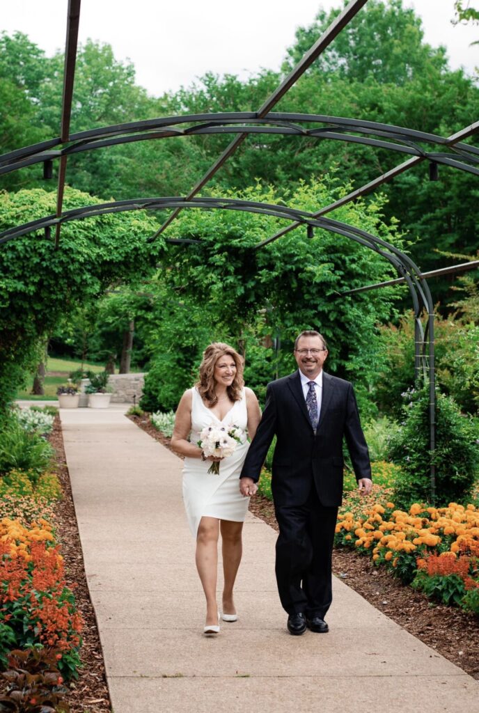 The bride and groom walk toward the camera under an arbor of greenery with orange and yellow flowers along the sides. The bride is wearing a fitted white dress with an asymmetrical hemline and plunging neckline. The groom is wearing a black suit with a white shirt and blue tie. The bride is holding a bouquet of white, ivory and blush flowers. They are at Cheekwood Botanical Garden in Nashville.