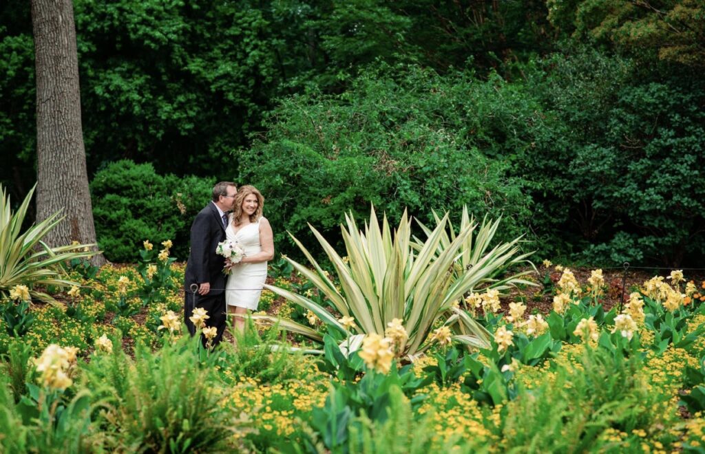 The bride and groom embrace in a lush garden full of greenery, palm fronds and orange and yellow flowers. The bride is wearing a fitted white dress with an asymmetrical hemline and plunging neckline. The groom is wearing a black suit with a white shirt and blue tie. The bride is holding a bouquet of white, ivory and blush flowers. The groom whispers into the bride's ear at Cheekwood Botanical Garden in Nashville.