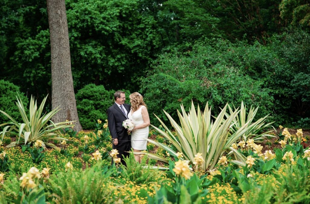 The bride and groom lean in for a kiss in a lush garden full of greenery, palm fronds and orange and yellow flowers. The bride is wearing a fitted white dress with an asymmetrical hemline and plunging neckline. The groom is wearing a black suit with a white shirt and blue tie. The bride is holding a bouquet of white, ivory and blush flowers. They are at Cheekwood Botanical Garden in Nashville.