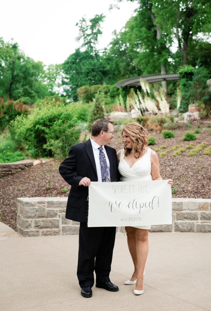 Groom wearing a black suit with a white shirt and blue tie smiles at the bride who is wearing a short form fitting white dress with a plunging neckline. They are holding a We Eloped sign at Cheekwood Botanical Gardens.
