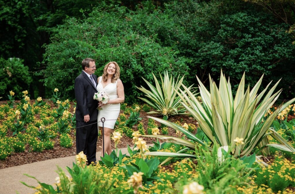 The bride and groom embrace and laugh in a lush garden full of greenery, palm fronds and orange and yellow flowers. The bride is wearing a fitted white dress with an asymmetrical hemline and plunging neckline. The groom is wearing a black suit with a white shirt and blue tie. The bride is holding a bouquet of white, ivory and blush flowers. They are at Cheekwood Botanical Garden in Nashville.