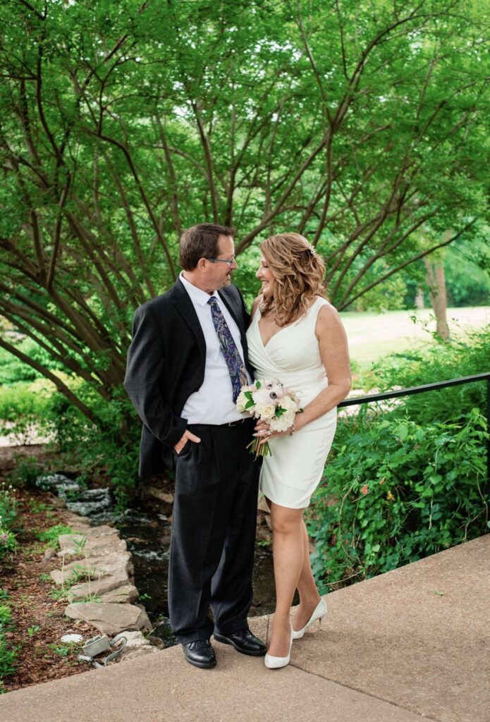 The bride and groom stand under a tree next to a stone path as they smile at each other. The bride is wearing a fitted white dress with an asymmetrical hemline and plunging neckline. The groom is wearing a black suit with a white shirt and blue tie. The bride is holding a bouquet of white, ivory and blush flowers. They are at Cheekwood Botanical Garden in Nashville.
