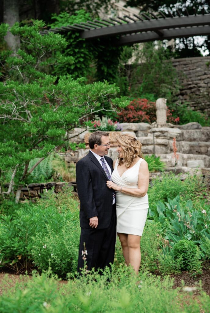The bride and groom embrace as they lean in for a kiss. The bride is wearing a fitted white dress with an asymmetrical hemline and plunging neckline. The groom is wearing a black suit with a white shirt and blue tie. They are standing in a garden with a stone wall behind them at Cheekwood Botanical Garden in Nashville.