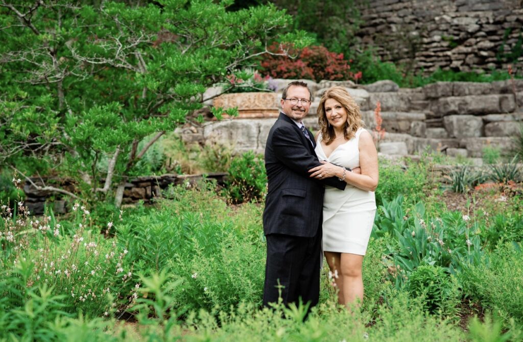 The bride and groom embrace in the garden. The bride is wearing a fitted white dress with an asymmetrical hemline and plunging neckline. The groom is wearing a black suit with a white shirt and blue tie. They are standing in a garden with a rough rock wall behind them at Cheekwood Botanical Garden in Nashville.