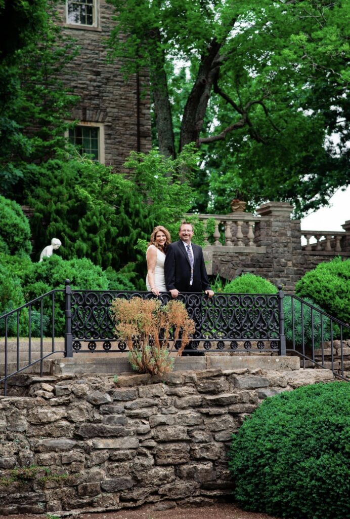 The bride and groom embrace as they over look the gardens at Cheekwood. They are standing at a filigreed iron work railing on a stone wall at Cheekwood Mansion. The bride is wearing a fitted white dress with a plunging neckline. The groom is wearing a black suit with a white shirt and blue tie. They are at Cheekwood Botanical Garden in Nashville.