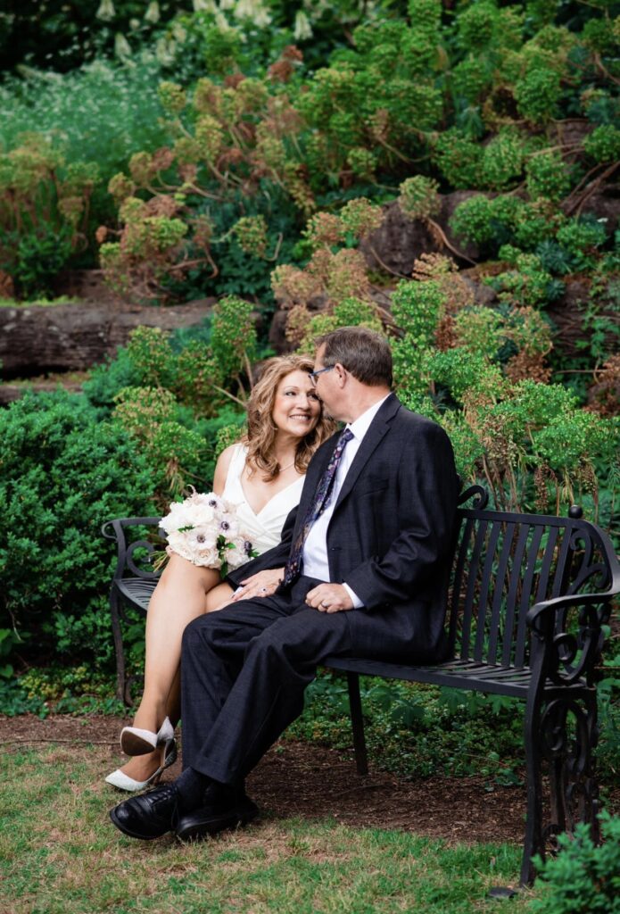 The bride and groom embrace as they lean in for a kiss. They are sitting on a black iron park bench. The bride is wearing a fitted white dress with a plunging neckline. The groom is wearing a black suit with a white shirt and blue tie. The bride is holding a bouquet of white, ivory and blush flowers. They are at Cheekwood Botanical Garden in Nashville.