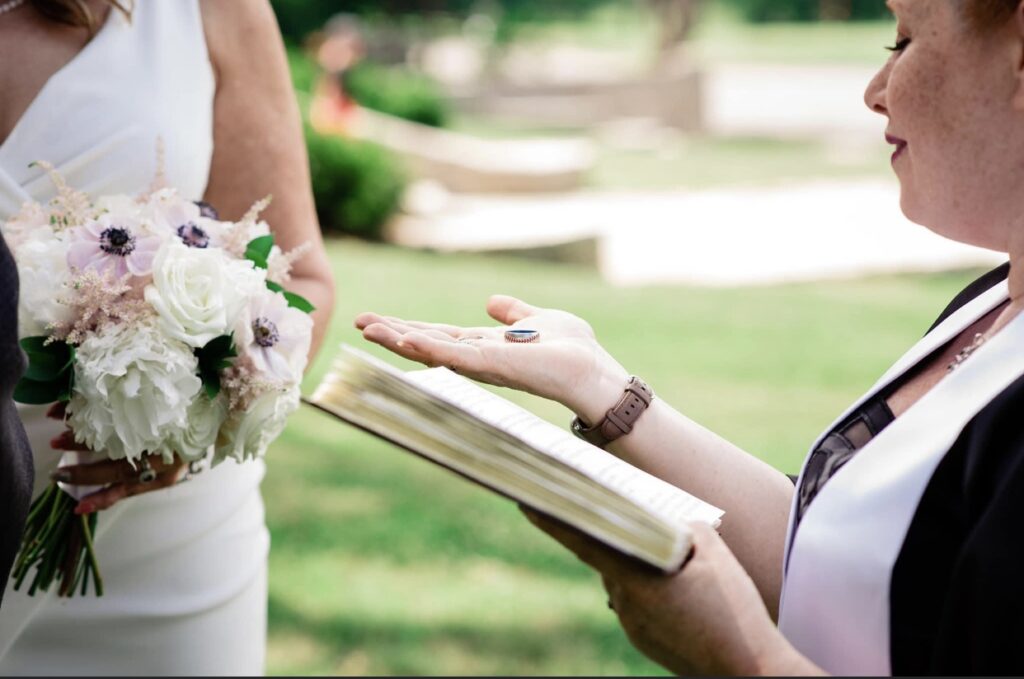 The wedding officiant, holding a book in one hand and wedding bands in the other, offer them to the bride. The bride is wearing a white dress with a plunging neckline. the bride is holding a bouquet of pink and white flowers. 