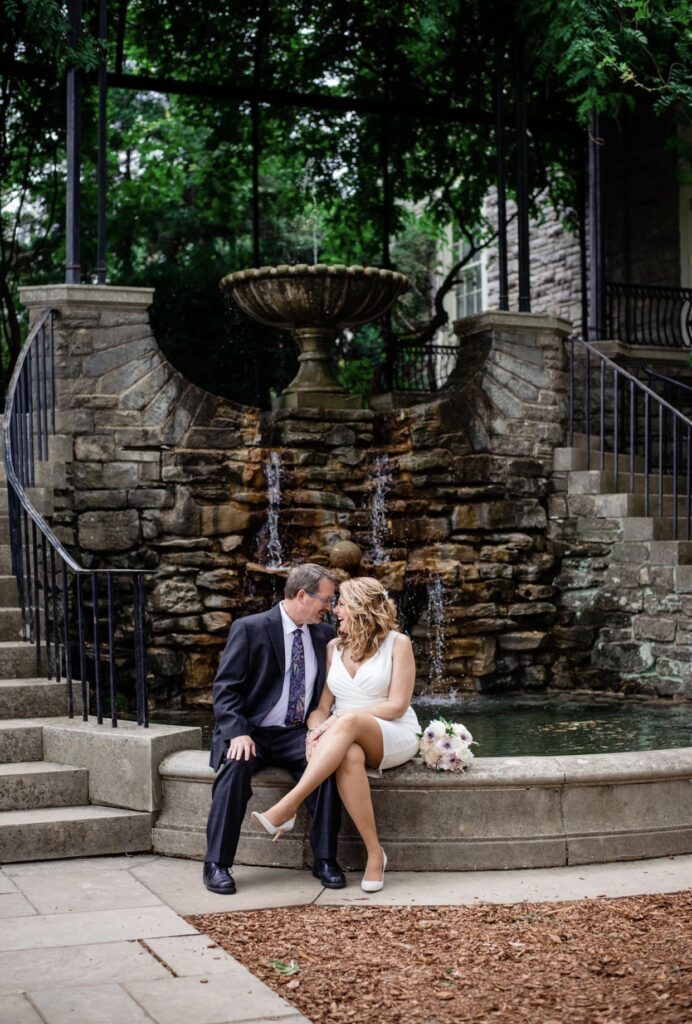 The bride and groom embrace as they lean in for a kiss. They are sitting in front of a waterfall fountain with spiraling staircases on either side. The bride is wearing a fitted white dress with an asymmetrical hemline and plunging neckline. The groom is wearing a black suit with a white shirt and blue tie. The bride's bouquet is sitting on the fountain next to her. It is a bouquet of white, ivory and blush flowers. They are at Cheekwood Botanical Garden in Nashville.