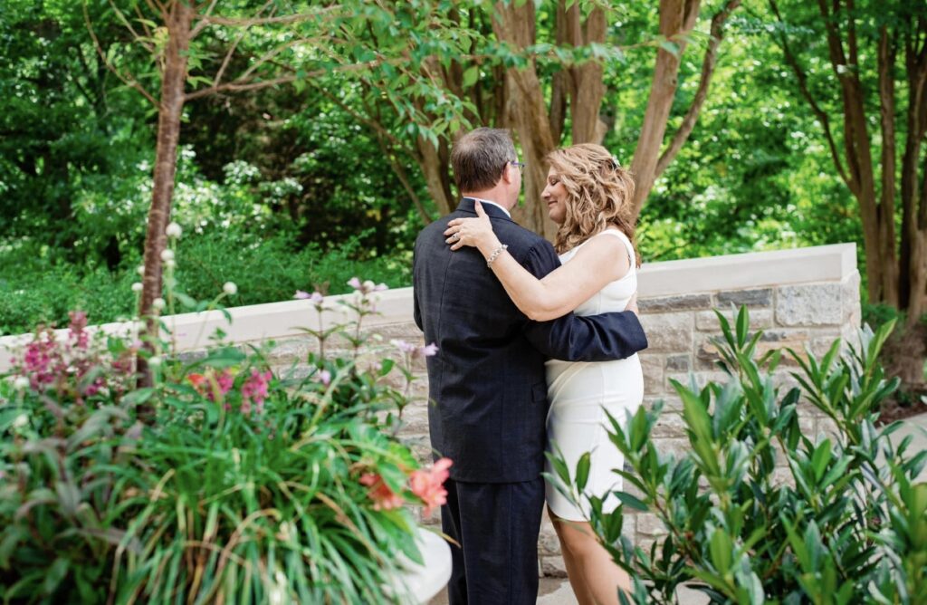 The bride and groom, looking away from the camera, dance  in a garden full of lush greenery, pink flowers and potted plants. The bride is wearing a short  fitted white dress. The groom is wearing a black suit. They are at Cheekwood Botanical Garden in Nashville.